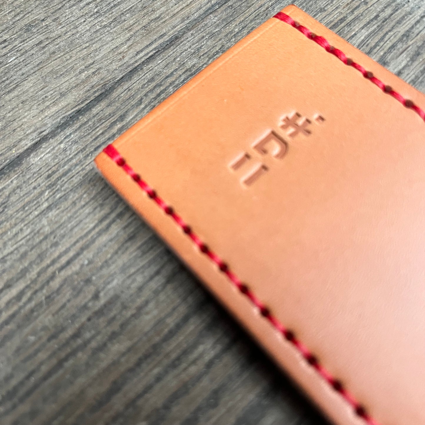 Eine Nahaufnahme der Niwaki-Lederscheide von Niwaki aus hellbraunem Leder mit roten Nähten und geprägten japanischen Katakana-Zeichen auf einem Holzhintergrund.