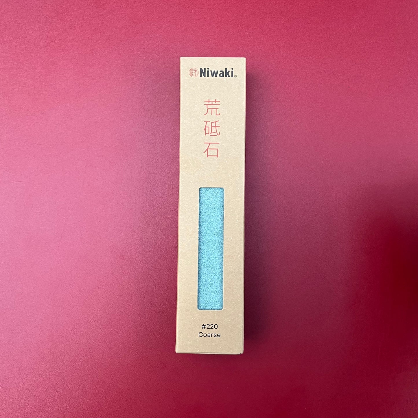 Der Niwaki-Schleifstein (#220 grob) von Niwaki wird in einer braunen Schachtel geliefert und steht vertikal auf einem roten Hintergrund – ideal zum Schärfen von Klingen und Gartengeräten.