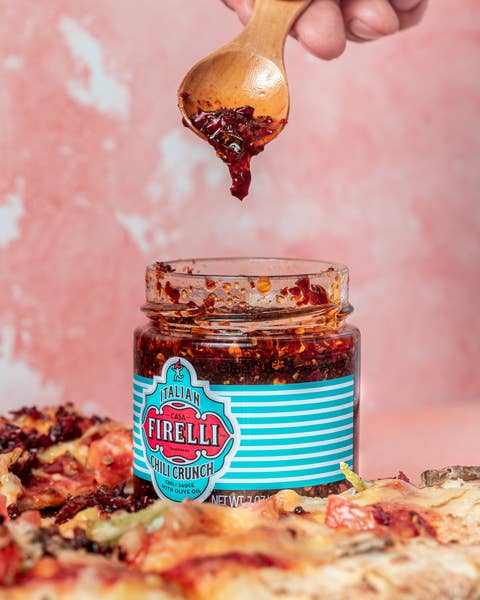 Eine Hand hält einen Holzlöffel mit Firelli | Chili Crunch 200g Glas über einem offenen Glas, hergestellt mit italienischem Olivenöl von Firelli. Unten ist eine mit würzigem Chili Crunch belegte Pizza zu sehen.