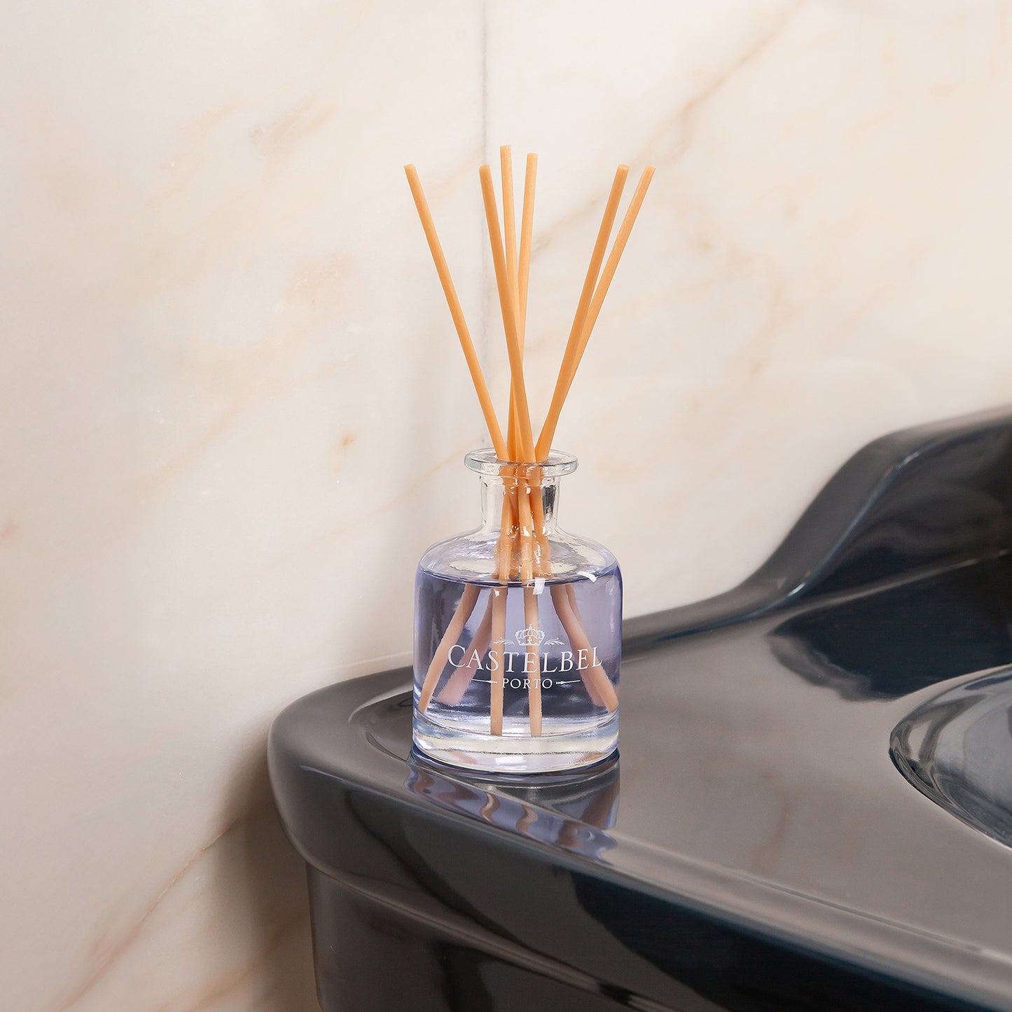 Ein Castelbel | Diffusor Lavendel 250 ml mit lila Flüssigkeit und Holzstäbchen steht auf einem dunklen Waschbecken und erfüllt die Luft vor einer hellen Marmorwand mit entspannendem Lavendelduft.