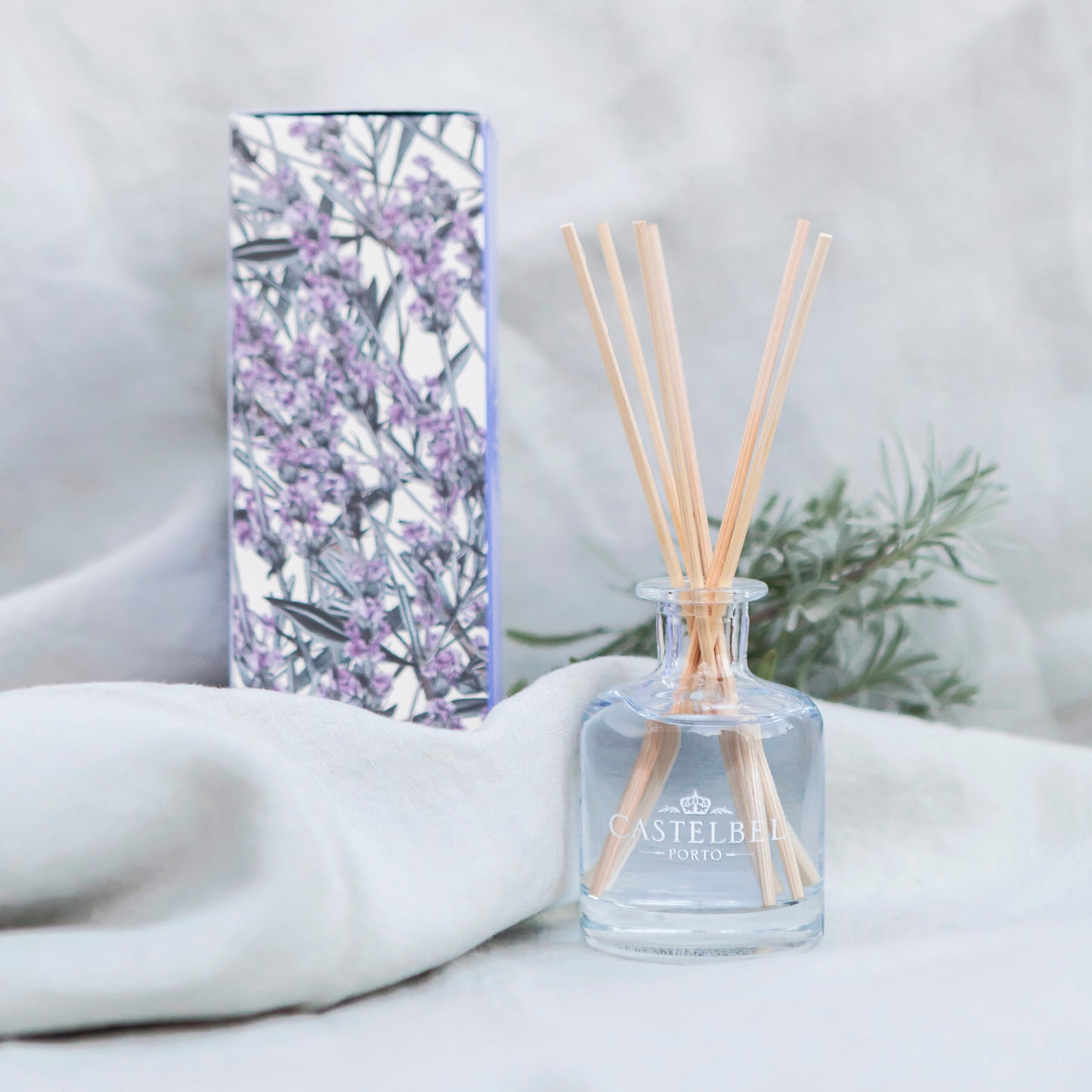 Ein Castelbel | Diffusor Lavendel 100 ml mit Holzstäbchen ruht auf einem weißen Tuch, begleitet von seiner Blumendose und Lavendelzweigen im Hintergrund, und erfüllt den Raum mit wohltuenden ätherischen Ölen.