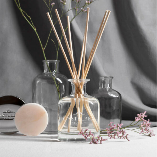 Der Castelbel | Diffusor White Jasmine 100 ml, ein klarer Glasdiffusor mit Holzblättern, sitzt zwischen Glasflaschen, einem beigen Schwamm und rosa Blüten auf hellgrauem Stoff. Dieser ölbasierte alkoholfreie Duft ist ideal für kleine Räume.