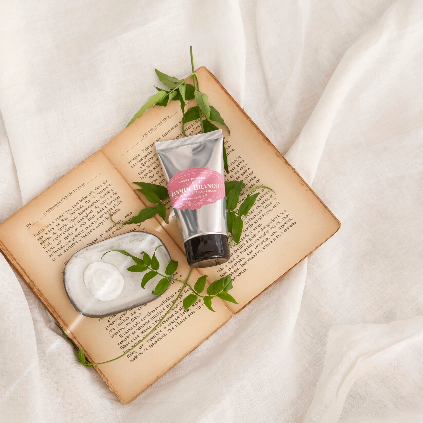 Eine Tube Castelbel | Handcreme Jasmin 60 ml und eine Seifenschale liegen auf einem aufgeschlagenen Buch mit verstreuten grünen Blättern; der blumige Duft schwebt über einem weichen weißen Stoff im Hintergrund.