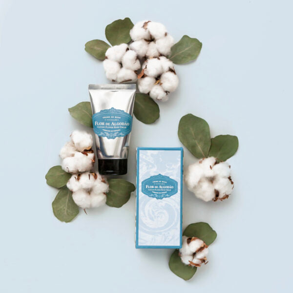 Eine silberne Tube Castelbel | Handcreme Cotton Flower 60 ml und eine blaue Schachtel ruhen auf einer hellblauen Fläche, umgeben von grünen Blättern und Baumwollblüten, die die pflanzlichen Buttersorten und Öle für intensive Feuchtigkeit hervorheben.