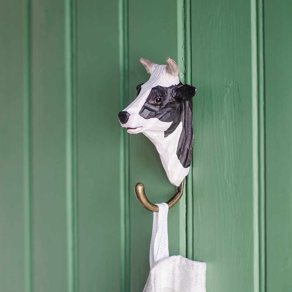 Der Wildlife Garden Handgeschnitzter Wandhaken aus Holz 'Kuh' von Wildlife Garden zeigt einen schwarz-weißen Kuhkopf und bronzefarbenen Metallhaken - ein originelles Geschenk für jedes Zuhause.