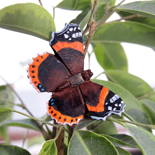 Ein 'Schmetterling Admiral' Magnet aus handgeschnitztem Holz von Wildlife Garden ruht auf grünen Blättern im Wildlife Garden und verschmilzt perfekt mit seiner Umgebung.