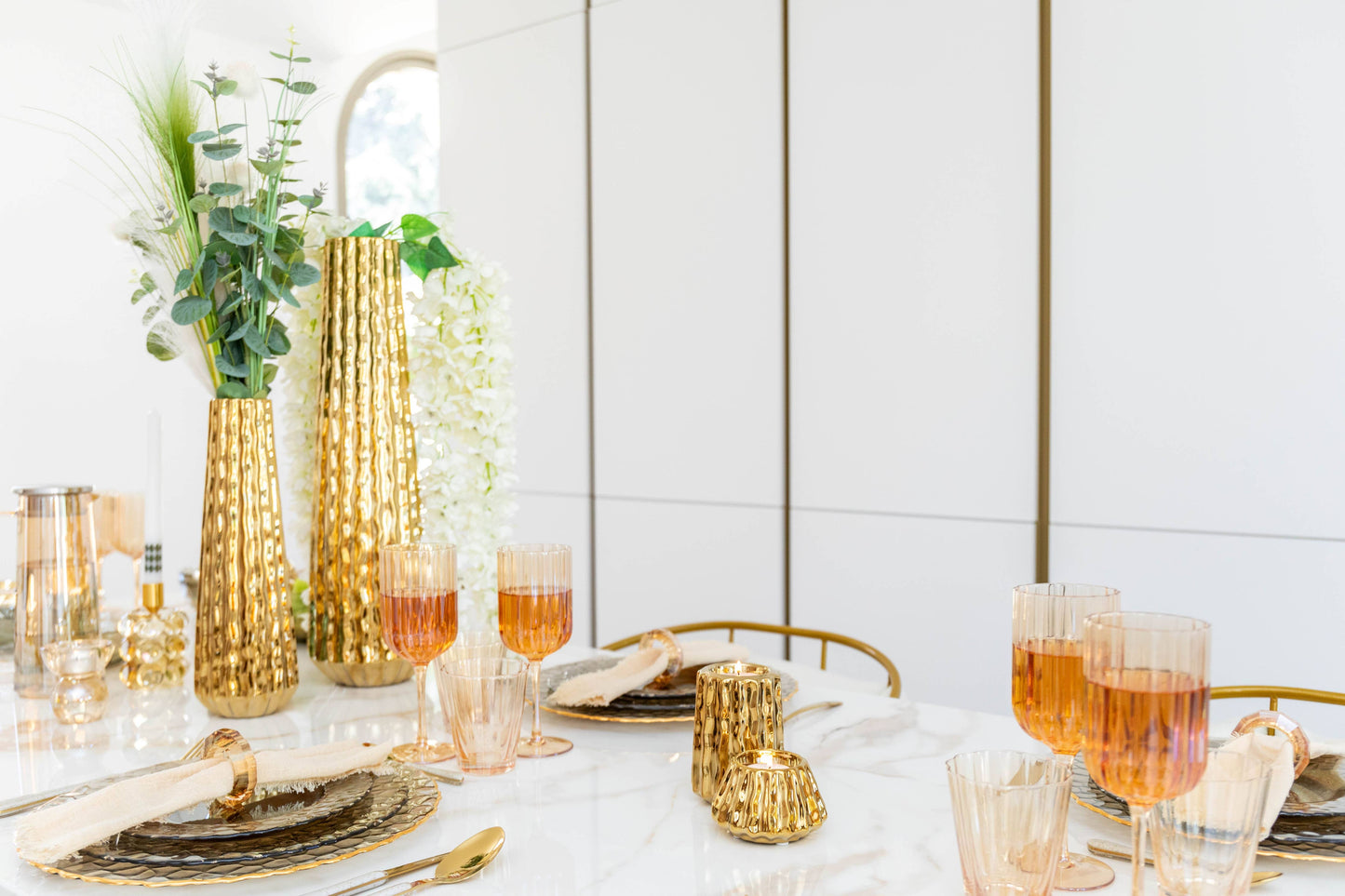 Ein modernes Esstisch-Set mit der goldenen Vase AULICA | Glänzende, goldenem Geschirr, erröteten Gläsern, cremefarbenen Servietten und grünem Laub auf einem weißen Marmortisch für einen eleganten und luxuriösen Look.