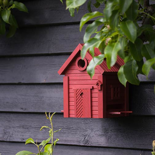 Der Wildlife Garden Nistkasten & Futterhaus "Red Earth" hat ein stilvolles Scheunendesign und ist an einer dunklen Holzwand montiert, umgeben von grünen Blättern und Zweigen.