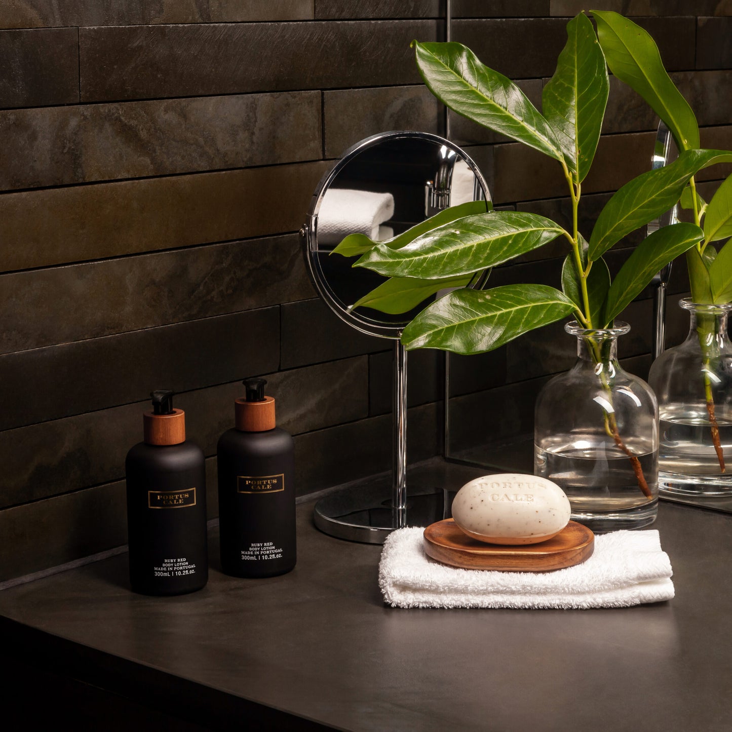 Ein moderner Badezimmertisch mit zwei schwarzen Seifenspendern, einem runden Spiegel, einem weißen Handtuch, Castelbel Body Lotion 'Rubinrot' Trauben & rote Beeren 300 ml, einem Stück Seife auf einer Holzschale und grünen Zweigen - alles vor dunkel gefliesten Wänden.