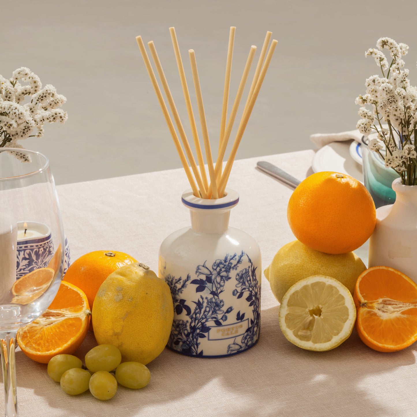 Der Castelbel Diffusor Gold & Blue mit rosa Pfeffer & Jasminduft (250 ml) steht auf einem Tisch mit Orangen, Zitronen, Trauben, weißen Blumen in Vasen, einem Weinglas und Tellern - alles auf einem hellen Tischtuch für ein elegantes Dufterlebnis.