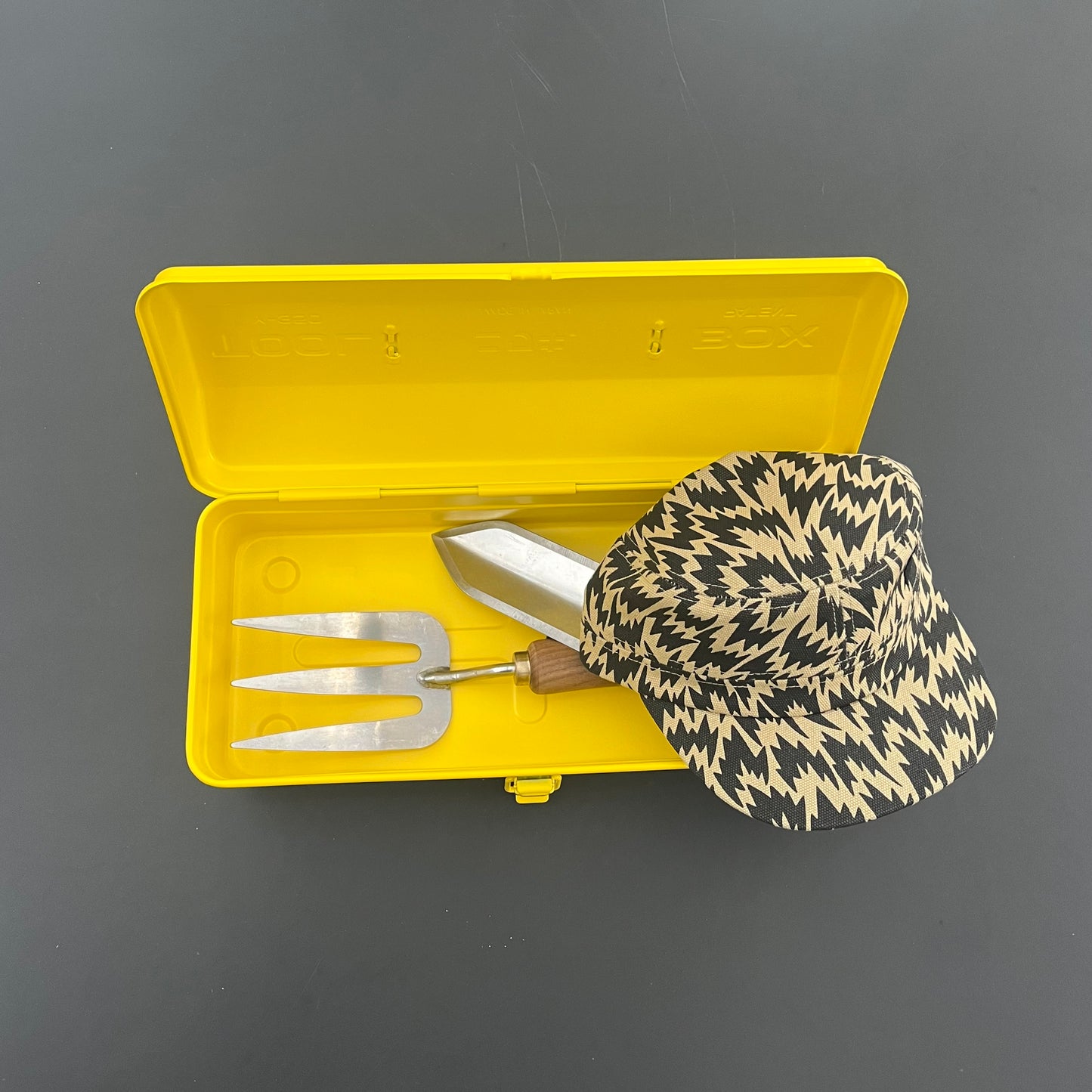 Die gelbe Niwaki Y-Type Toolbox von Niwaki enthält eine handgeführte Gartengabel und eine Kelle, gekrönt von einem schwarz-gelben Zickzack-Hut, alles auf einer dunkelgrauen Oberfläche - ideal, um Ihre Gartenutensilien zu organisieren.