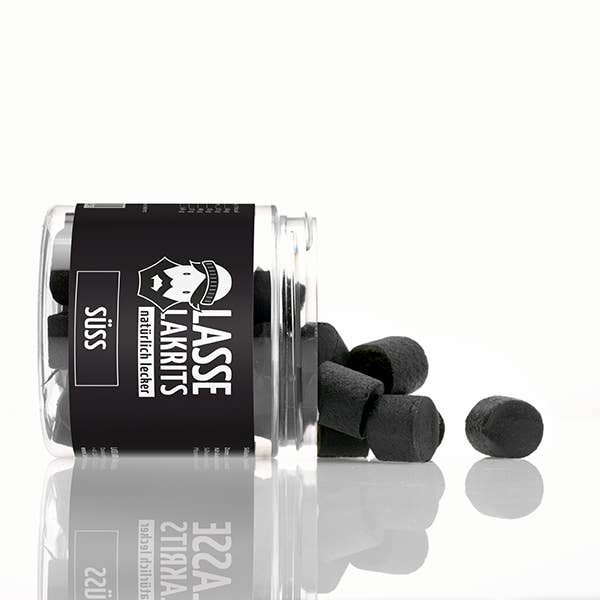Ein Glasgefäß mit der Aufschrift LASSE LAKRITS | Dose Lakritz Süss natürlich lecker liegt auf der Seite und verschüttet veganes Lakritz auf eine weiße, reflektierende Oberfläche. Auf dem schwarzen Etikett steht der Markenname LASSE LAKRITS in weißer Schrift und Grafik.