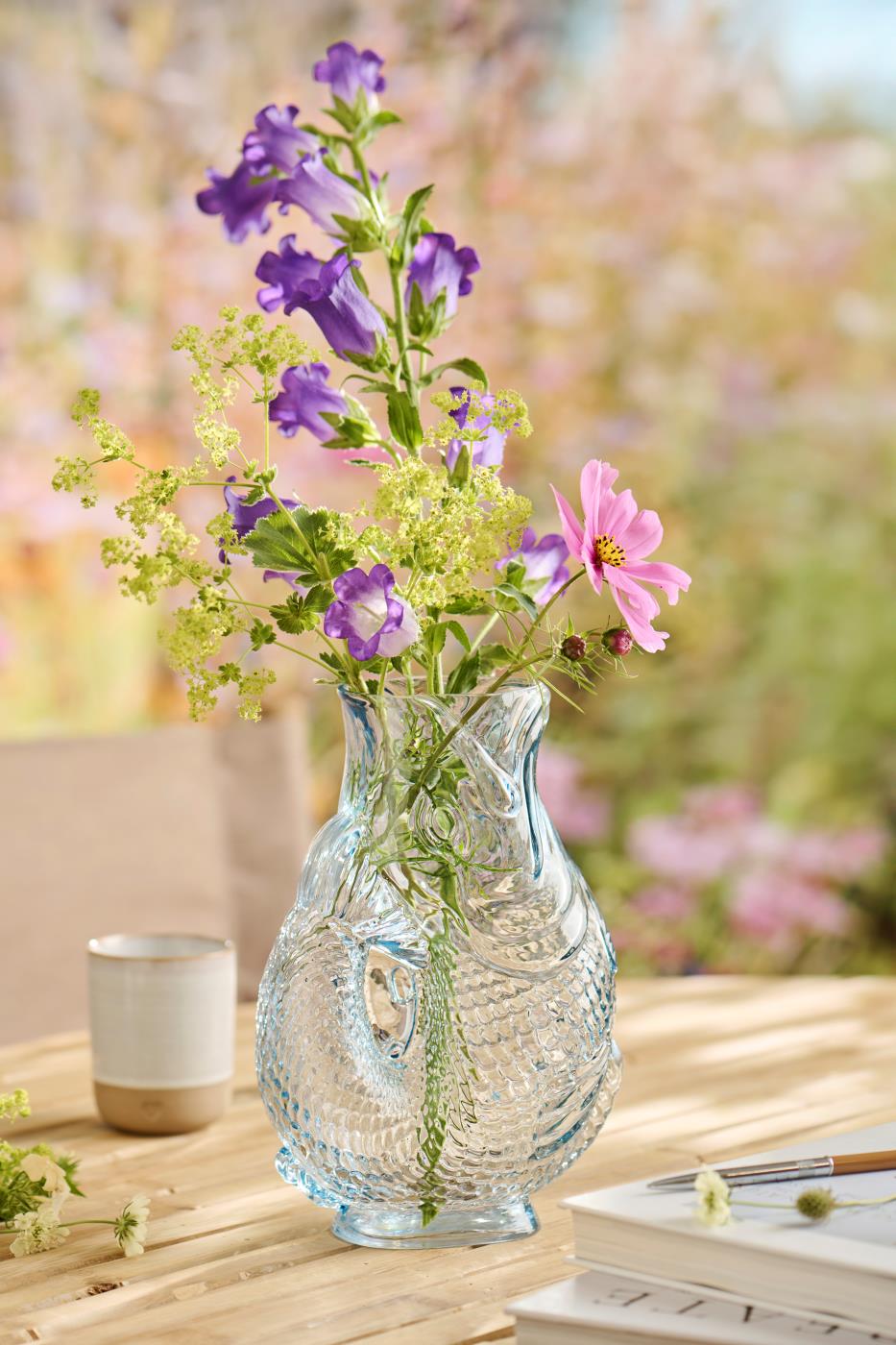 Die Ib Laursen Vase fischförmig in Hellblau, 24 cm hoch von Königliche Gartenakademie, präsentiert lila, pinke und grüne Blumen auf einem Holztisch mit einer Tasse und verschwommenem Garten im Hintergrund.