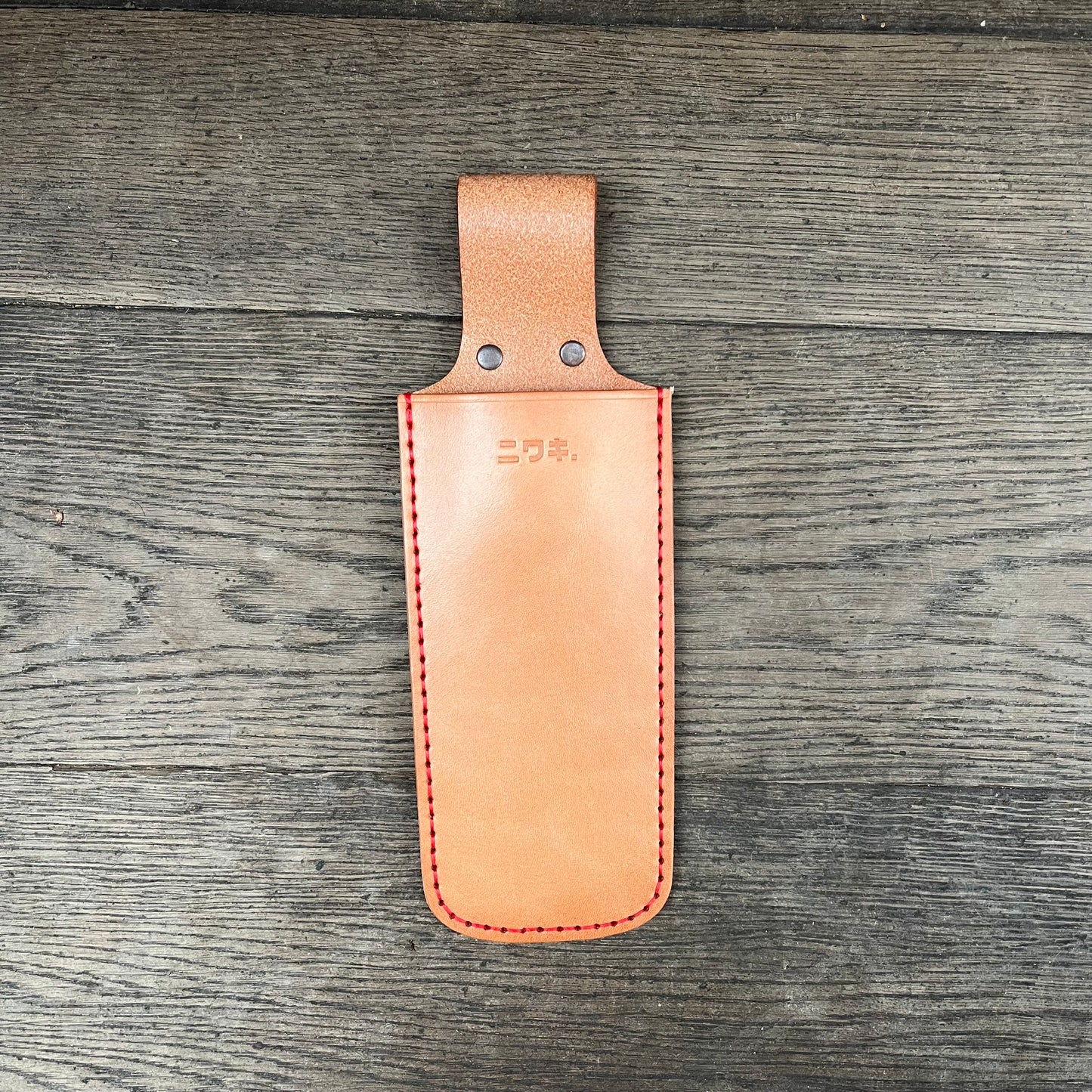 Eine hellbraune Niwaki | 'Saw Holster' hochwertige Lederschutzhülle Klappsäge aus Leder mit roten Nähten und zwei Metallnieten liegt flach auf Holz. Sie hat eine Gürtelschlaufe und ein Niwaki-Logo, ideal zur Sicherung Ihrer Klappsäge.