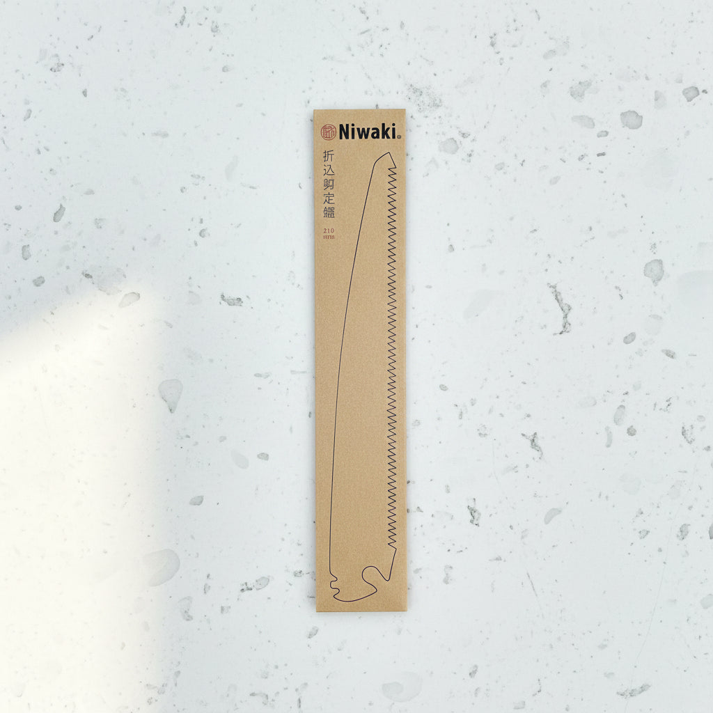 Eine rechteckige Pappverpackung mit der Aufschrift Niwaki, auf der die Niwaki | Ersatzklinge für Niwaki Gartenschere GR 210 abgebildet ist, mit einer Linienillustration, japanischen Schriftzeichen und "210" auf einer Holzoberfläche.