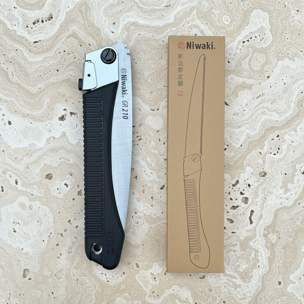 Eine Niwaki 'GR 210 Folding Saw' Klappsäge mit schwarzem Griff und silbernem Sägeblatt wird neben ihrer braunen Schachtel gezeigt, die eine Outdoor Säge Strichzeichnung zeigt, alles vor einem kräftigen roten Hintergrund.