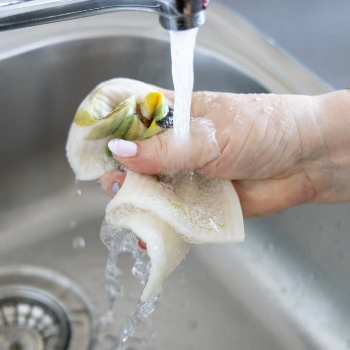 Eine Hand spült das More Joy Home | 'Orangen' biologisch abbaubares Schwammtuch unter fließendem Wasser aus einer Küchenarmatur, die nachhaltige Reinigung über einer Edelstahlspüle präsentiert.