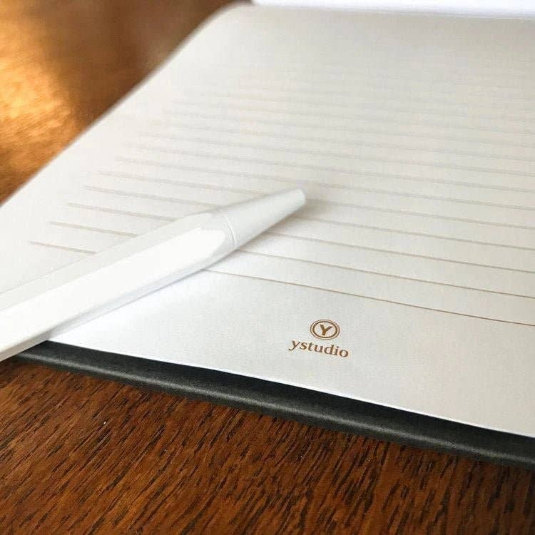 Ein weißer Stift liegt auf einem aufgeschlagenen, linierten ENTREPÔT YSTUDIO | Klassisches Reflect-Briefpapier-Set, mit "ystudio" am Seitenende, das an hochwertiges Kopierbriefpapier erinnert und elegant auf einer Holzunterlage liegt.