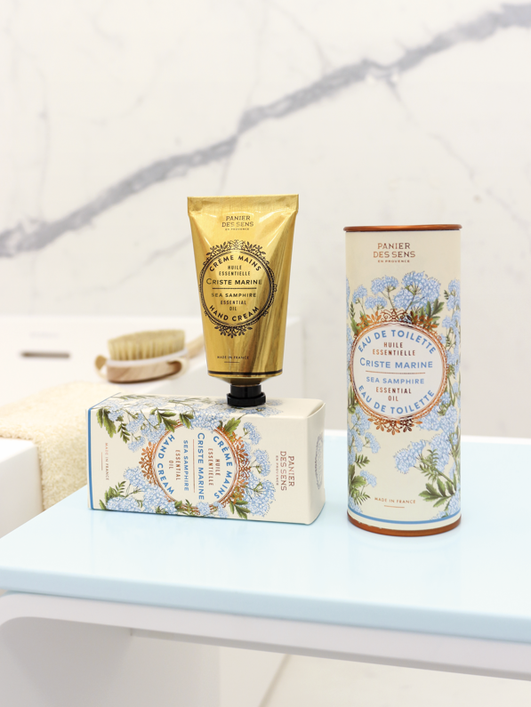 Eine goldene Handcremetube und eine Schachtel Panier Des Sens Pflanzliche Seife Meerfenchel 150 g stehen auf einer hellblauen Fläche mit einer Marmorwand und einer Bürste im Hintergrund.