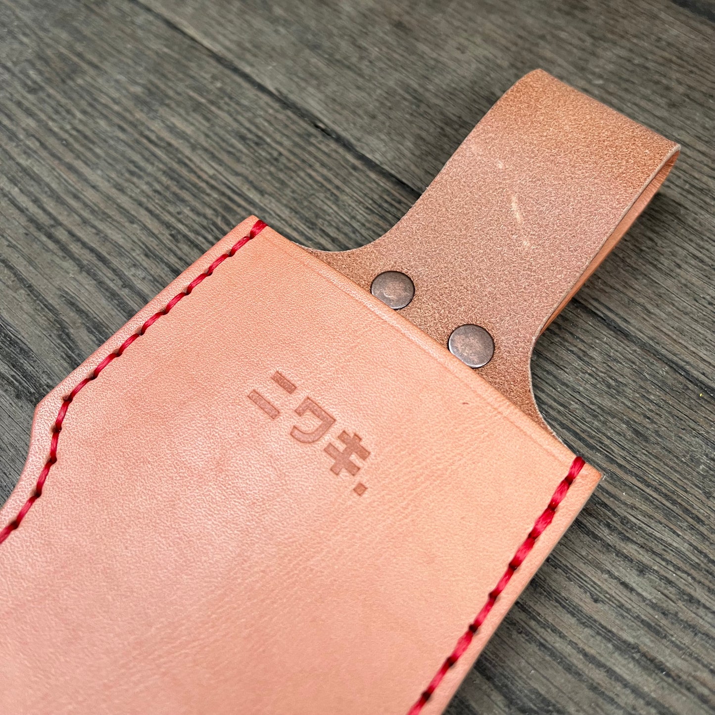 Eine Nahaufnahme der Niwaki | 'Single Holster' Lederschutzhülle für Schere, aus hellbraunem Leder mit roten Nähten, zwei Metallnieten und eingeprägten japanischen Schriftzeichen, dargestellt auf einem hölzernen Hintergrund.