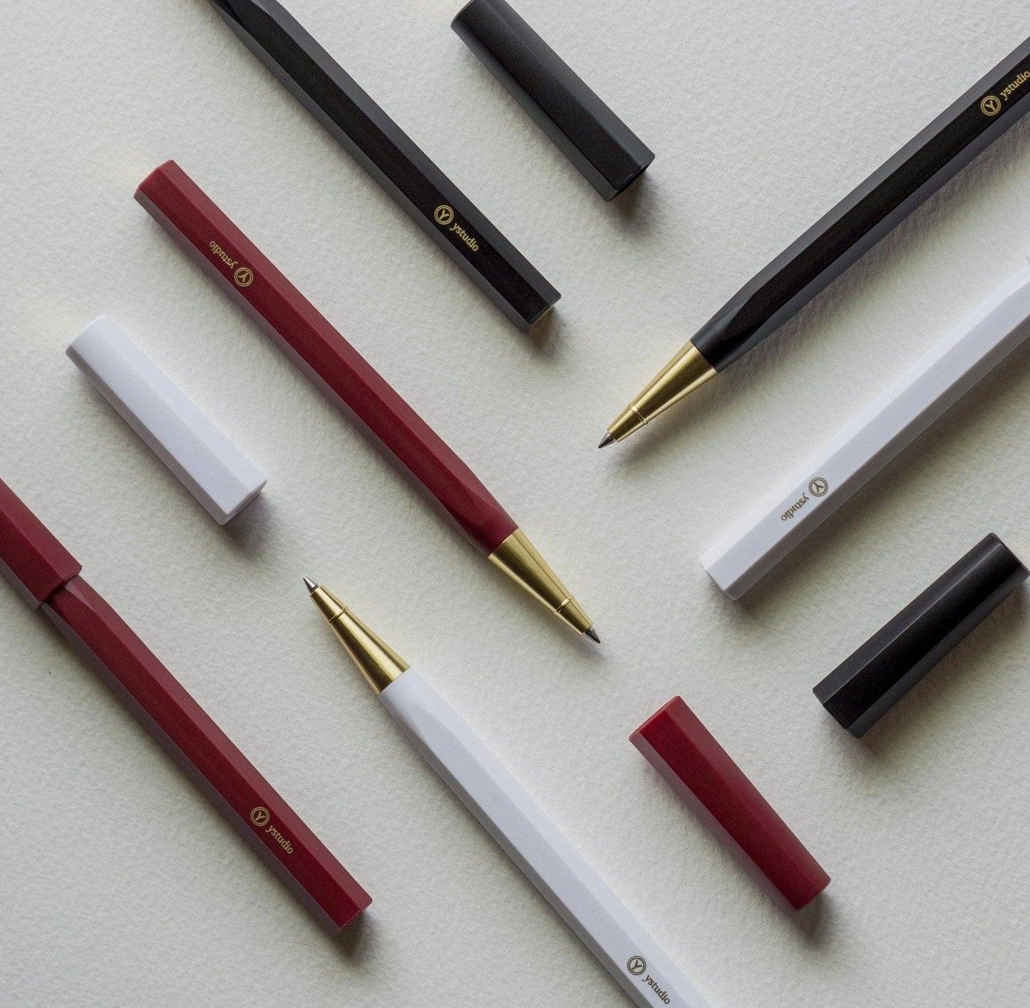 Die ENTREPÔT YSTUDIO Harz-Rollerball-Stifte (Weiß) mit Acryl- und Messingkorpus, Goldspitzen und passenden Kappen stehen diagonal auf einer strukturierten weißen Fläche.