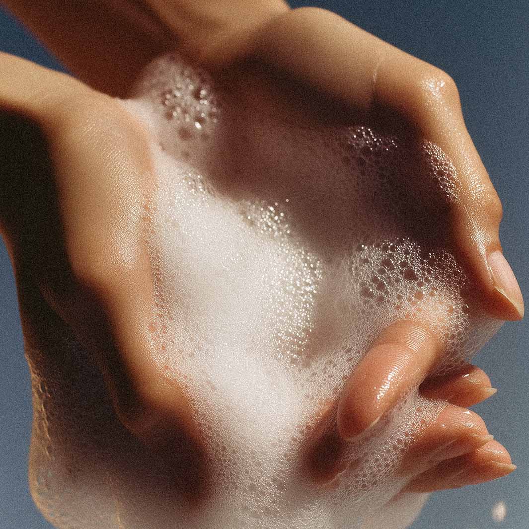 Nahaufnahme von zwei Händen, die sanft weißen, seifigen Schaum mit sichtbaren Blasen schöpfen, der die Hautstruktur und den Schaum von Panier Des Sens | Duftseife Rose des Bastides 150 g vor einem weichen, unscharfen Hintergrund hervorhebt.