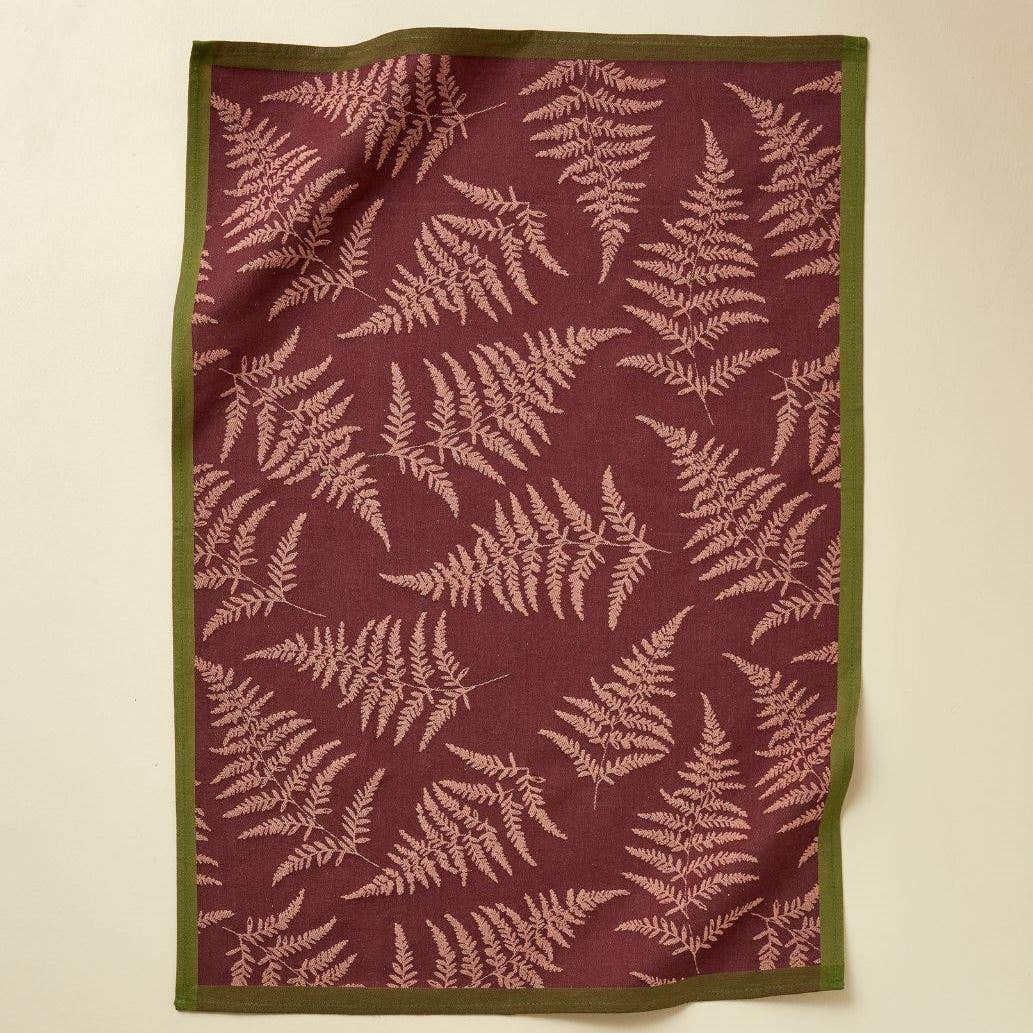 Das Sylvie Thiriez Jacquard Geschirrtuch 'Verzauberter Wald' in Bordeaux ist ein rechteckiges Baumwollgeschirrtuch mit kastanienbraunem Hintergrund, hellrosa Farnmotiv und grüner Bordüre, das flach auf einer hellen Oberfläche liegt.