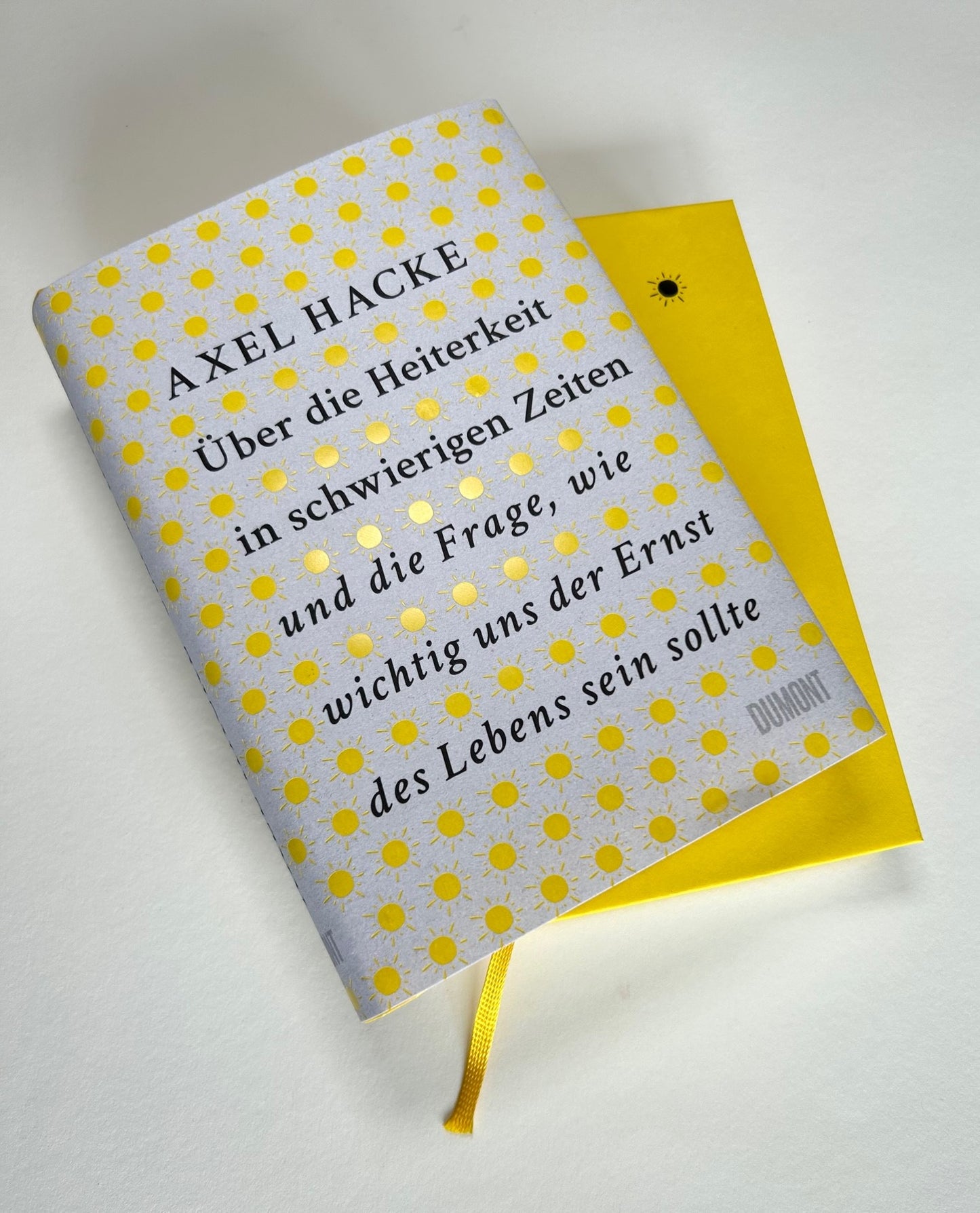 Das deutsche Buch "Über die Heiterkeit | Unterhaltsam, klug und persönlich" aus dem Dumont Verlag zeigt ein sonnengemustertes Cover auf leuchtend gelbem Hintergrund, inspiriert von Friedrich Schillers Konzept der Heiterkeit.