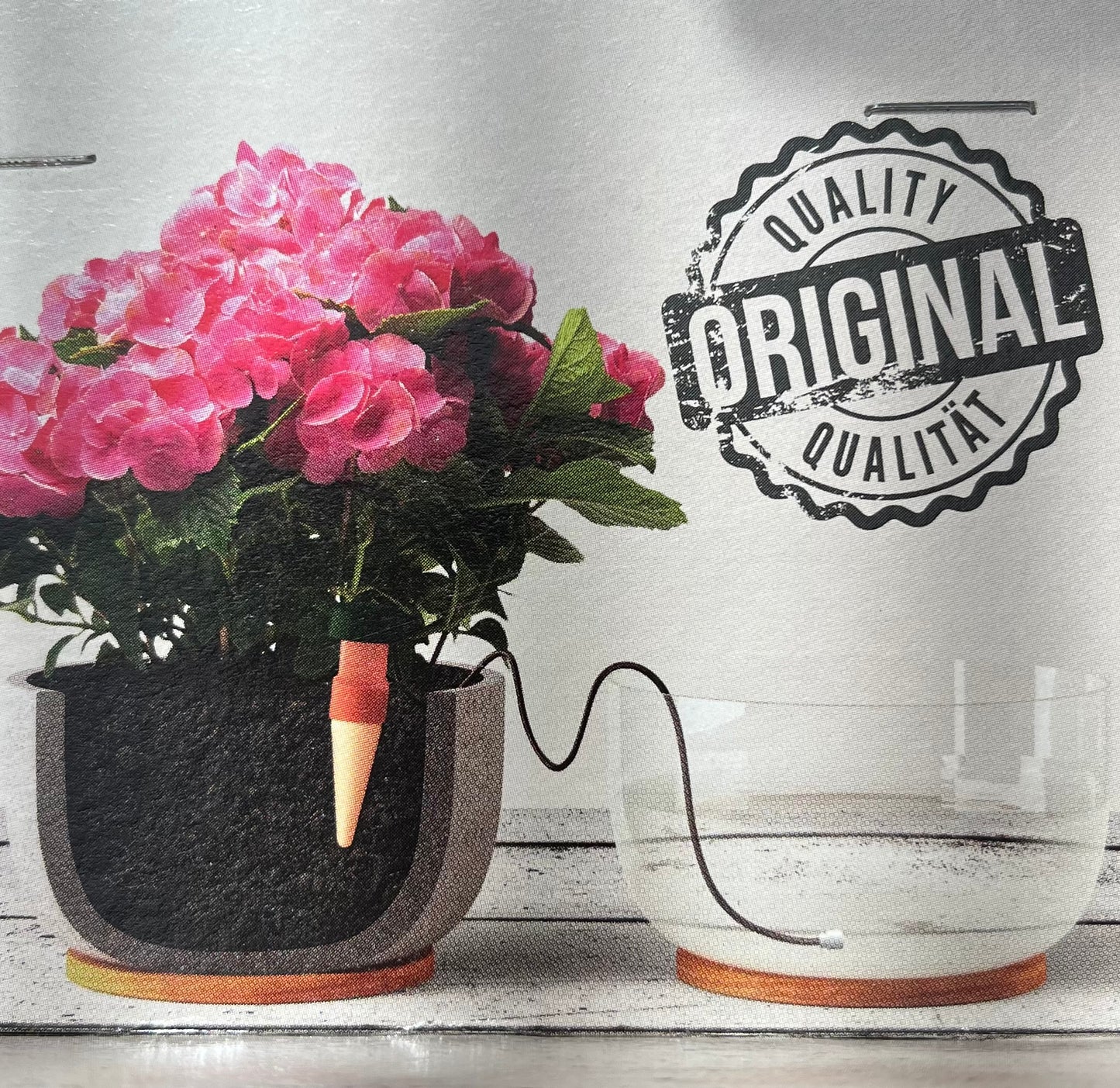 A Blumat | 'Classic' Praktische Bewässerung für Zimmer- und Balkonpflanzen versorgt eine Pflanze mit pinken Blüten über einen Schlauch aus einem transparenten Wasserbehälter. Im Bild ist oben rechts das Original Blumat Qualitätssiegel zu sehen.