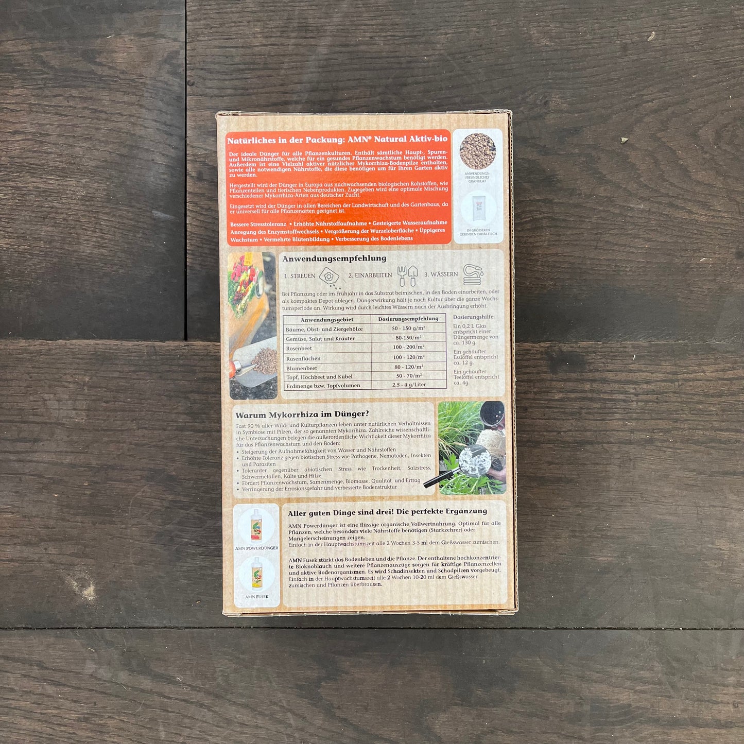 Auf einem dunklen Holzboden steht eine Kartonverpackung mit der Aufschrift „AMN Natural Aktiv-bio“ von AMN, mit deutschem Text, Dosierungsanweisungen und Pflanzenbildern. Die Schachtel beschreibt gesunde Düngung und Mykorrhiza für den natürlichen Gartenbau.