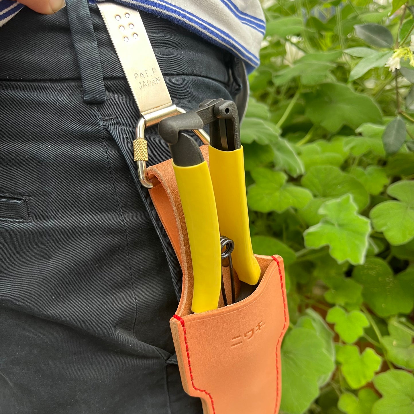 Eine Person in dunkler Hose befestigt die Niwaki 'Belt Clip' Praktische Halterung für Niwaki Single Holster von Niwaki an ihrer Tasche, während sie ein Gartengerät mit gelbem Griff und grünem Laub im Hintergrund hält.