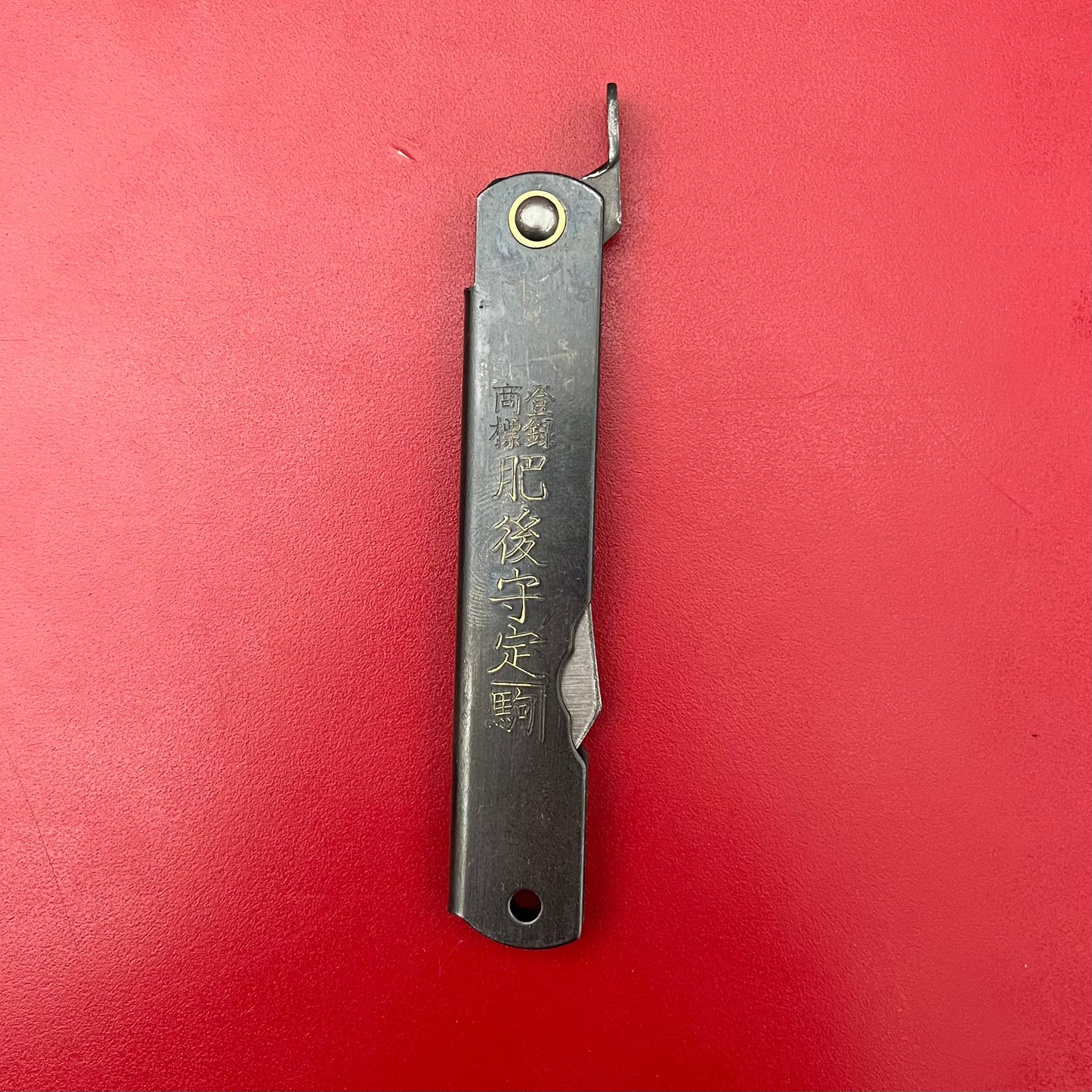 Das Niwaki | 'SK Higonokami Folding Knife' Klappmesser der Marke Niwaki mit Kanji-Gravur auf dem Metallgriff liegt geschlossen auf leuchtend rotem Hintergrund.