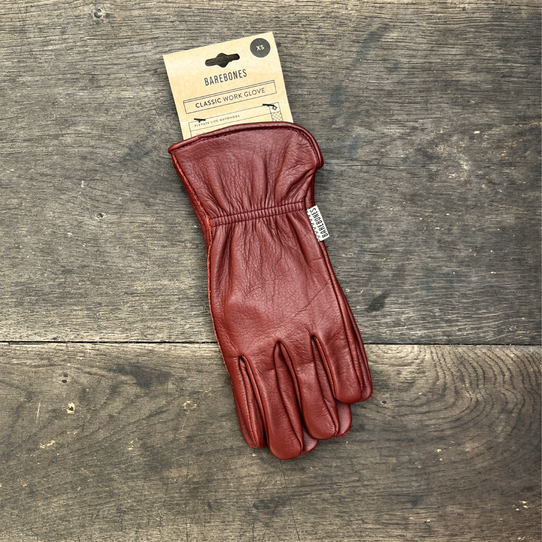 Ein einzelner cognacfarbener Gartenhandschuh aus Leder im Stil der „Barebones“ | Gartenhandschuhe aus Büffelleder „Cognac“ von der Königlichen Gartenakademie liegt auf einer verwitterten Holzoberfläche.