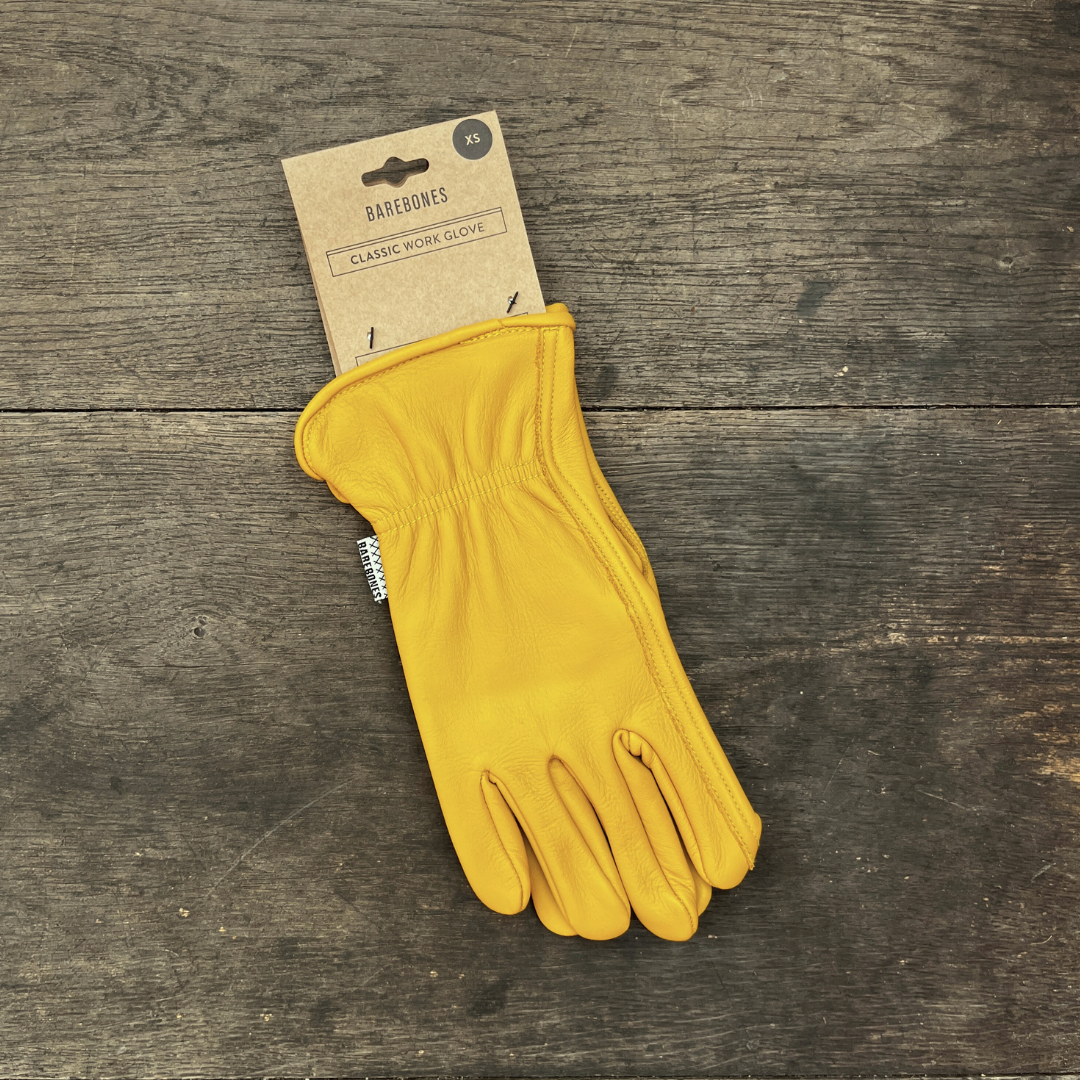 Ein Paar „Barebones“ | Gartenhandschuhe aus Büffelleder „Natural“ in Größe XS von der Königlichen Gartenakademie, versehen mit einem Kartonetikett mit der Aufschrift „Barebones Classic Work Glove“, liegt auf einer rustikalen Holzfläche.