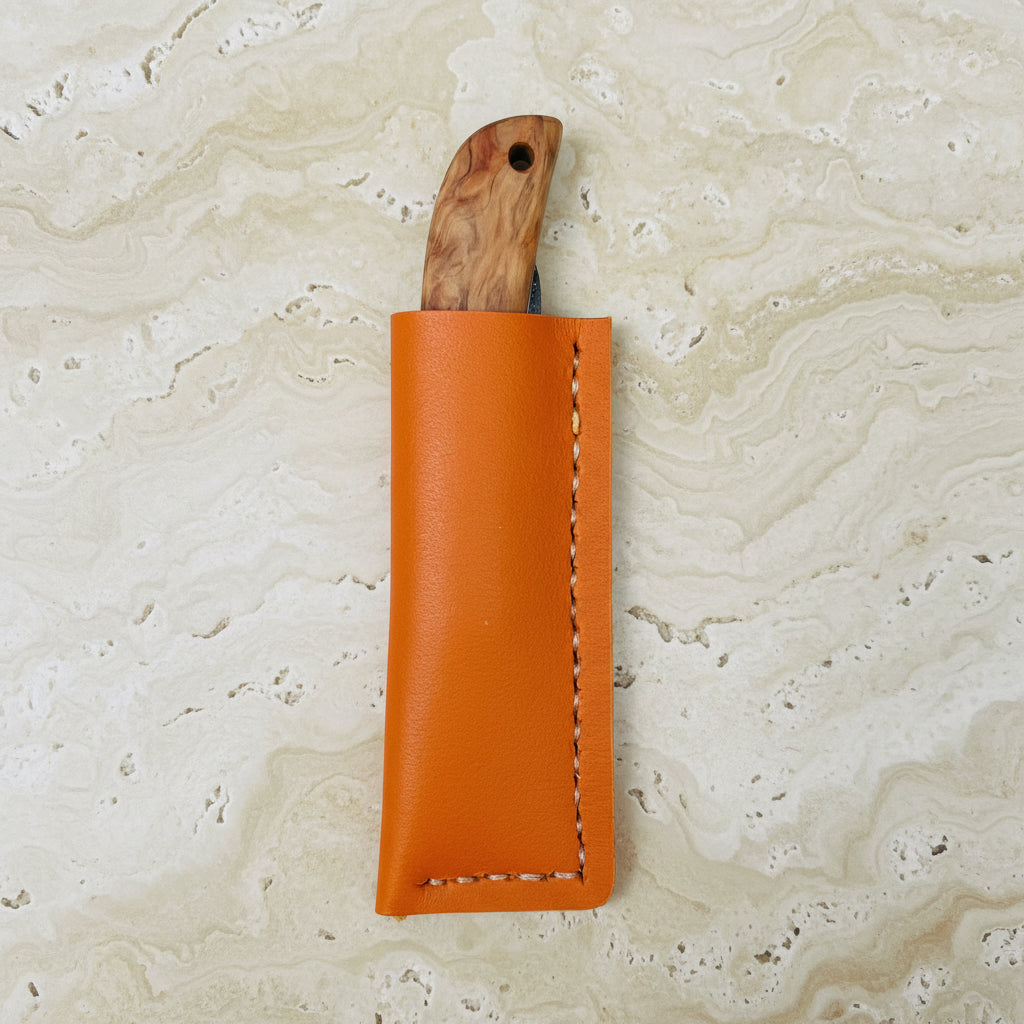 Das 'Kotoh X Niwaki Knife' Taschenmesser von Niwaki mit Holzgriff liegt teils in seiner orangefarbenen Lederschutzhülle auf Holz - ideal für Gartenarbeit oder als zuverlässiges Alltagsmesser.