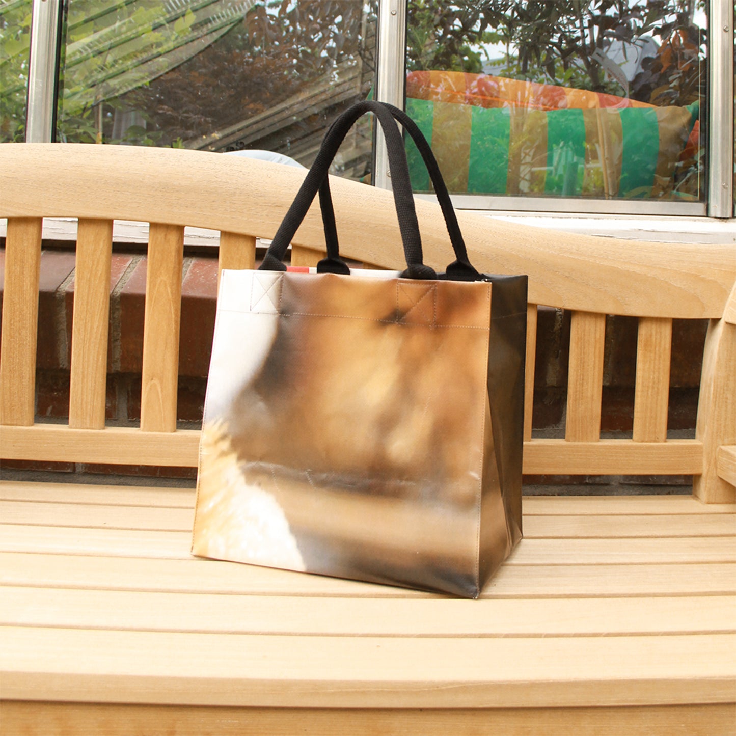 Die Upcycling Tasche 'Torf' aus Werbeplane von Königliche Gartenakademie, in Braun und Beige mit schwarzen Griffen, ruht auf einer hellen Holzbank im Freien, eingerahmt von einem Fenster und bunten Kissen.