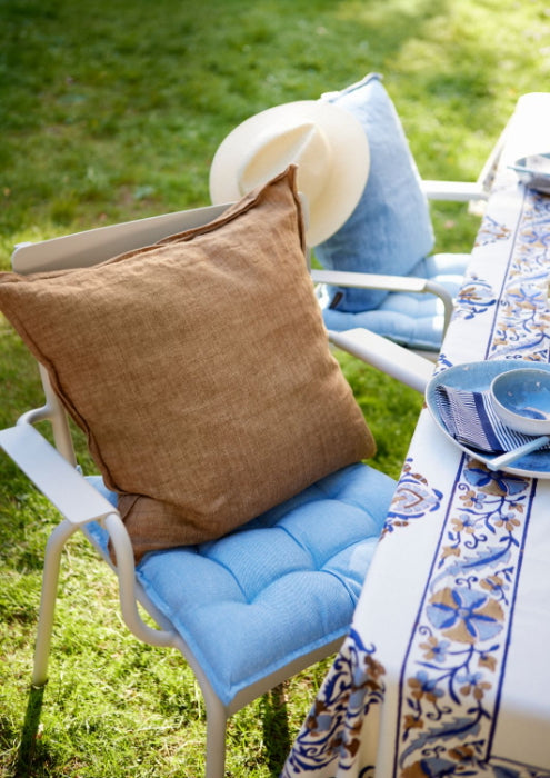 Zwei weiße Terrassenstühle mit blauen Kissen und einem Bungalow 'Milan Ocean Blue' Sitzkissen 45 x 45 cm stehen an einem Tisch mit einer blau-weißen Tischdecke. Ein Stuhl hat ein braunes Kissen und einen Strohhut auf seiner Rückenlehne, alles auf grünem Gras im Freien platziert.