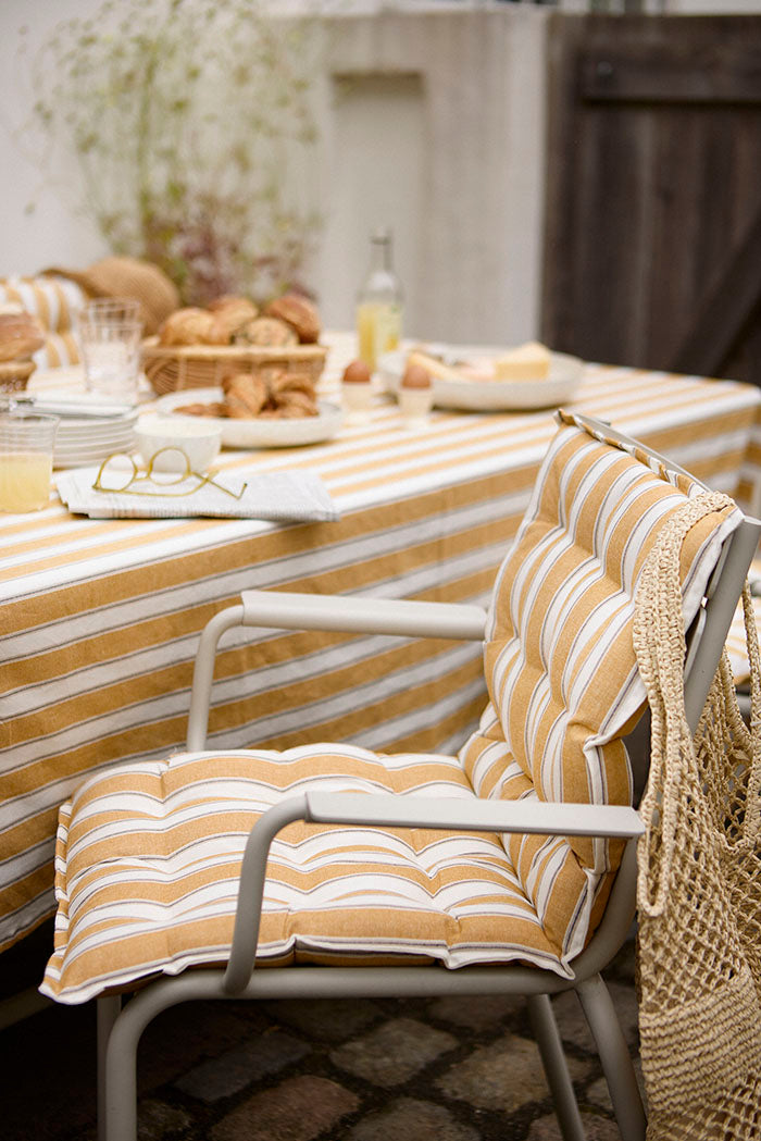 Ein gemütlicher Esstisch im Freien ist mit dem gelb-weiß gestreiften Sitzkissen 'Palermo Honey' von Bungalow im Format 45 x 90 cm ausgestattet. Frisches Brot, Getränke, Gläser und eine Strohtasche auf dem Stuhl sorgen für eine entspannte, einladende Atmosphäre.