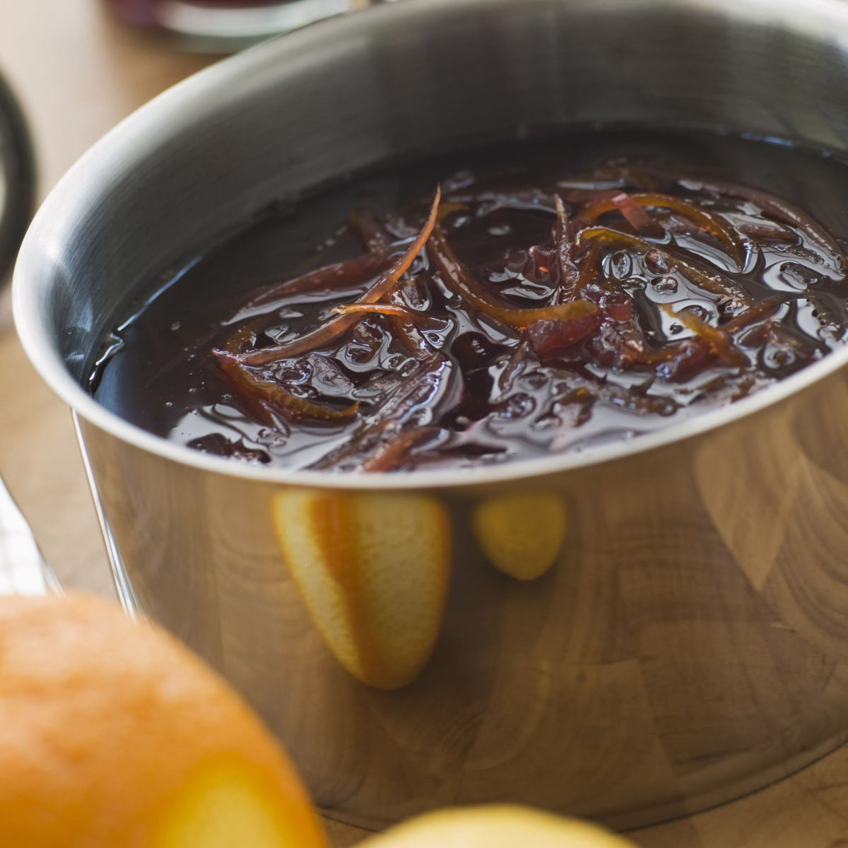 Ein Edelstahltopf ist bis zum Rand gefüllt mit glänzender, dunkelbrauner „Wilkin & Sons | Tiptree Cumberland Sauce“ (227 g), deren Oberfläche mit Zitrusschalenstreifen verziert ist. Die Szene, die auf einer Holzunterlage ruht, wird durch verschwommene Orangen und Zitronen im Vordergrund vervollständigt. Diese fruchtig-würzige Sauce, die von Wilkin & Sons nach der Tradition ihrer klassischen Rezepte hergestellt wird, besticht durch ein reichhaltiges Aroma und eine leuchtende Farbe.