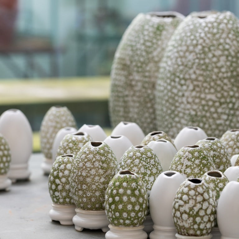 Eine Reihe kleiner Des Pots | Vase Ei-auf-Schale Form (H 10 cm, Ø 5,5 cm) der Königlichen Gartenakademie - einige schlicht weiß, andere mit grüner und weißer Blumenglasur - sind ausgestellt, dahinter zwei größere gemusterte Vasen.