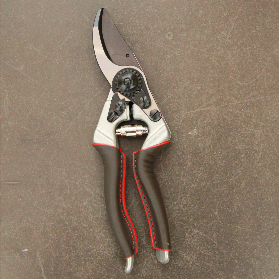 Ein Paar FELCO 6 Elite - Kompakte Gartenschere von Felco mit braunen Griffen und roten Nähten ruht auf einer braunen Oberfläche, mit einer glänzenden Metallklinge und Getriebemechanismus für präzise Schnitte in der Nähe des Drehpunkts.