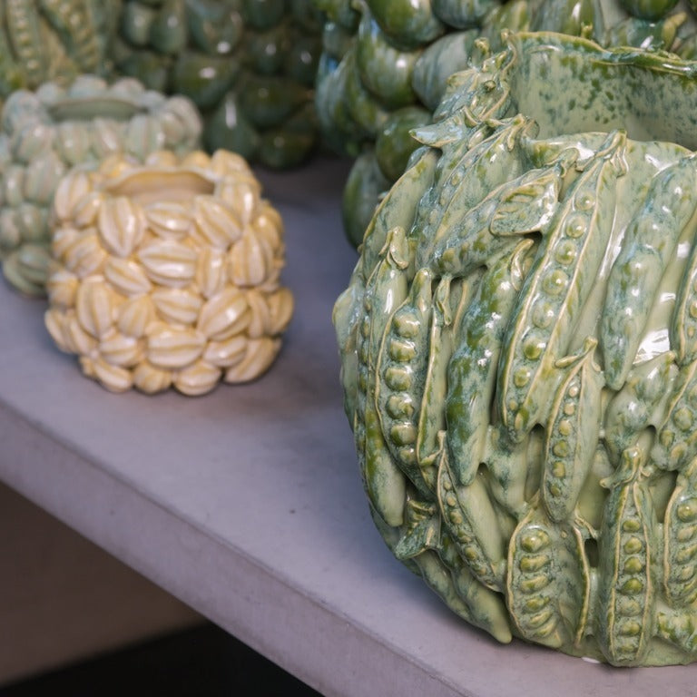 Zwei grüne Des Pots Keramikvasen sitzen auf einer Oberfläche: Die vordere Vase "Des Pots | Vase grün mit Schoten" zeigt glänzende Erbsenschoten, die kleinere dahinter leuchtende Pistazienschalen. Beide bieten eine organische Textur als Deko.