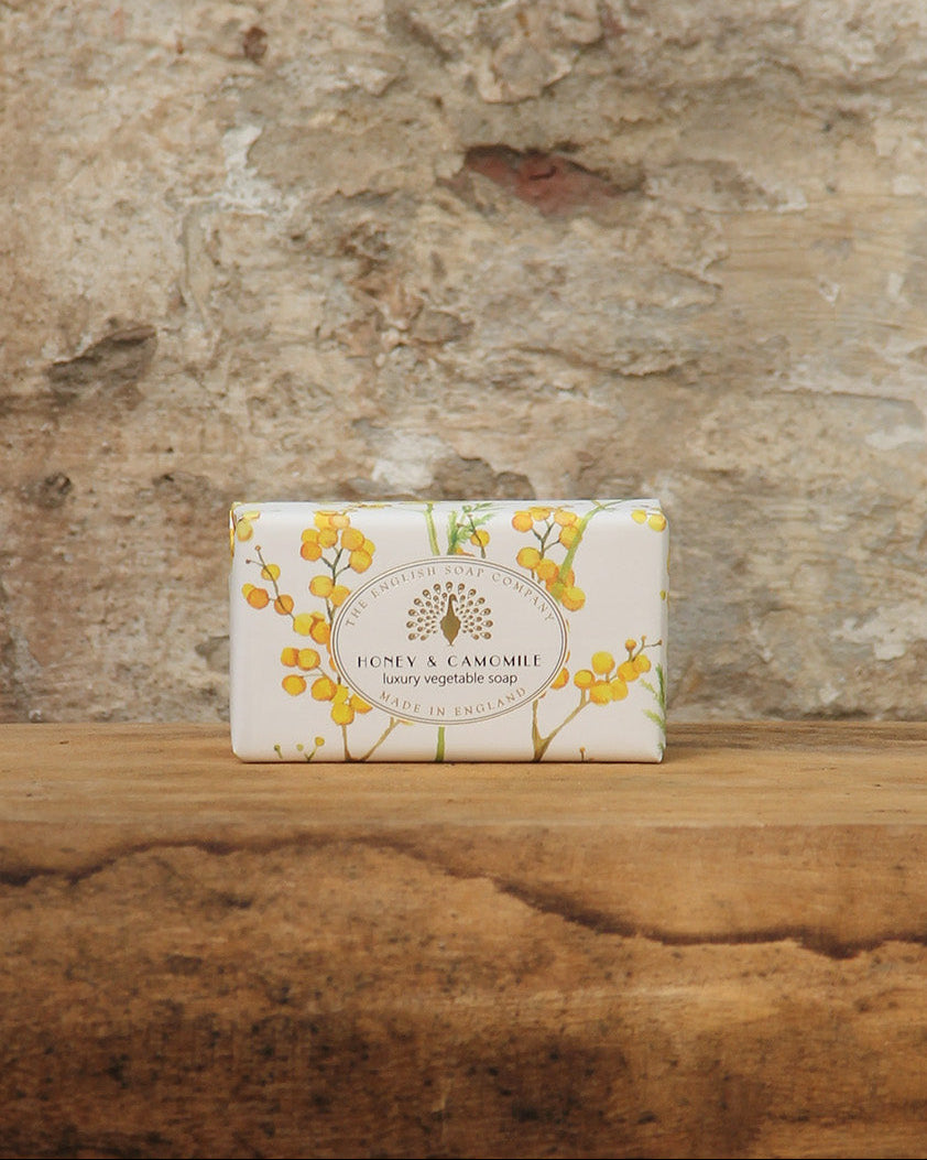 Ein Stück pflanzliche Handseife mit fruchtigem Honig-und Kamillenduft von The English Soap Company ESC, eingewickelt in Blumenpapier, ruht auf rustikalem Holz vor einer strukturierten Steinwand.