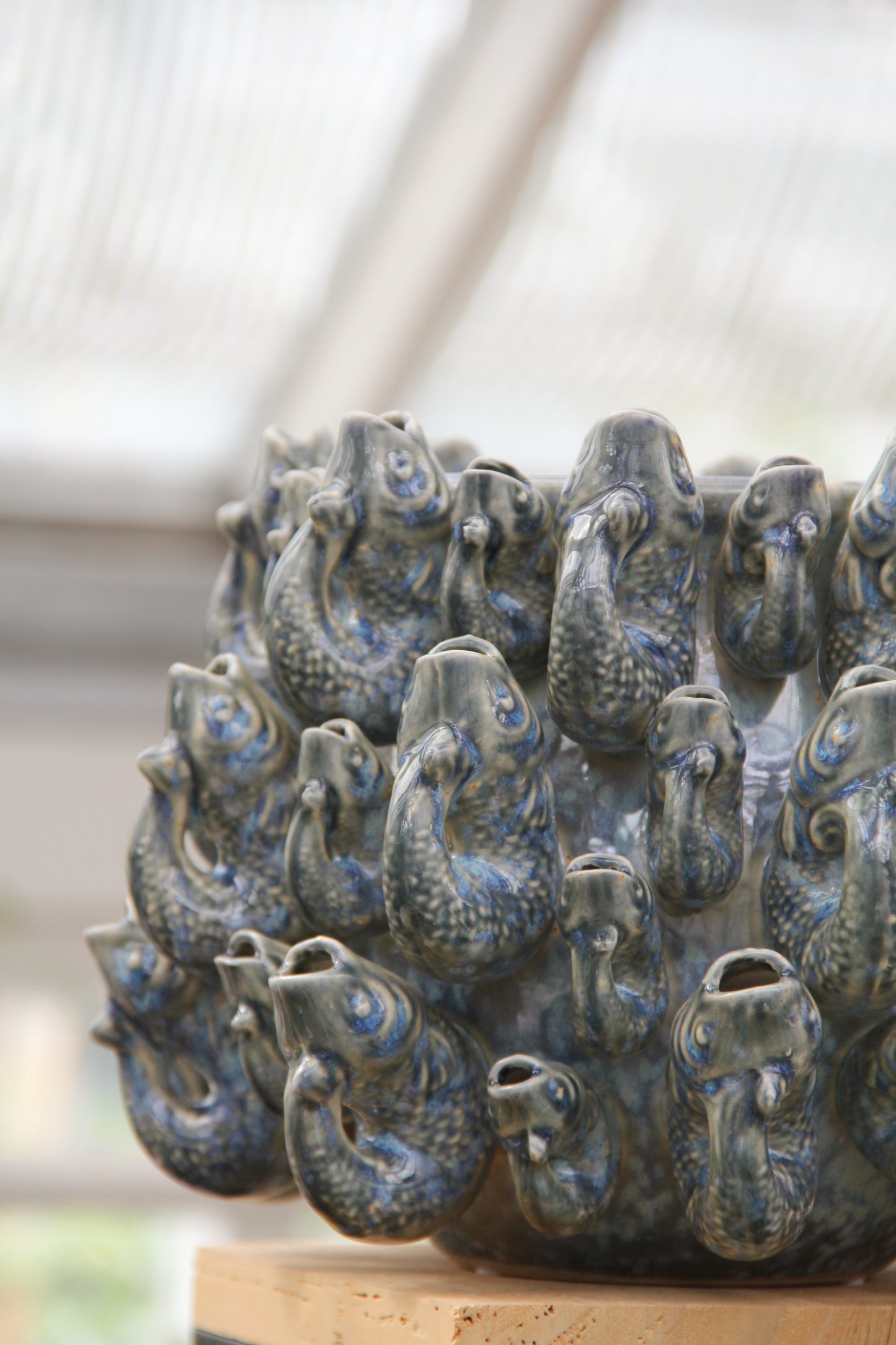 Wir stellen vor: Des Pots | Vase „Fisch“, klein, in Blau aus Steingut (H 18 cm, Ø 23 cm) von Des Pots – ein handgeformtes Designstück aus Steingut, glasiert in Blautönen, das mit mehreren aneinandergereihten Fischköpfen und geöffneten Mündern beeindruckt. Diese außergewöhnliche Designer-Vase verbindet kunstvolle Handwerkskunst mit markanter optischer Ausstrahlung und wird stilvoll auf einem Holzsockel im Innenbereich präsentiert.