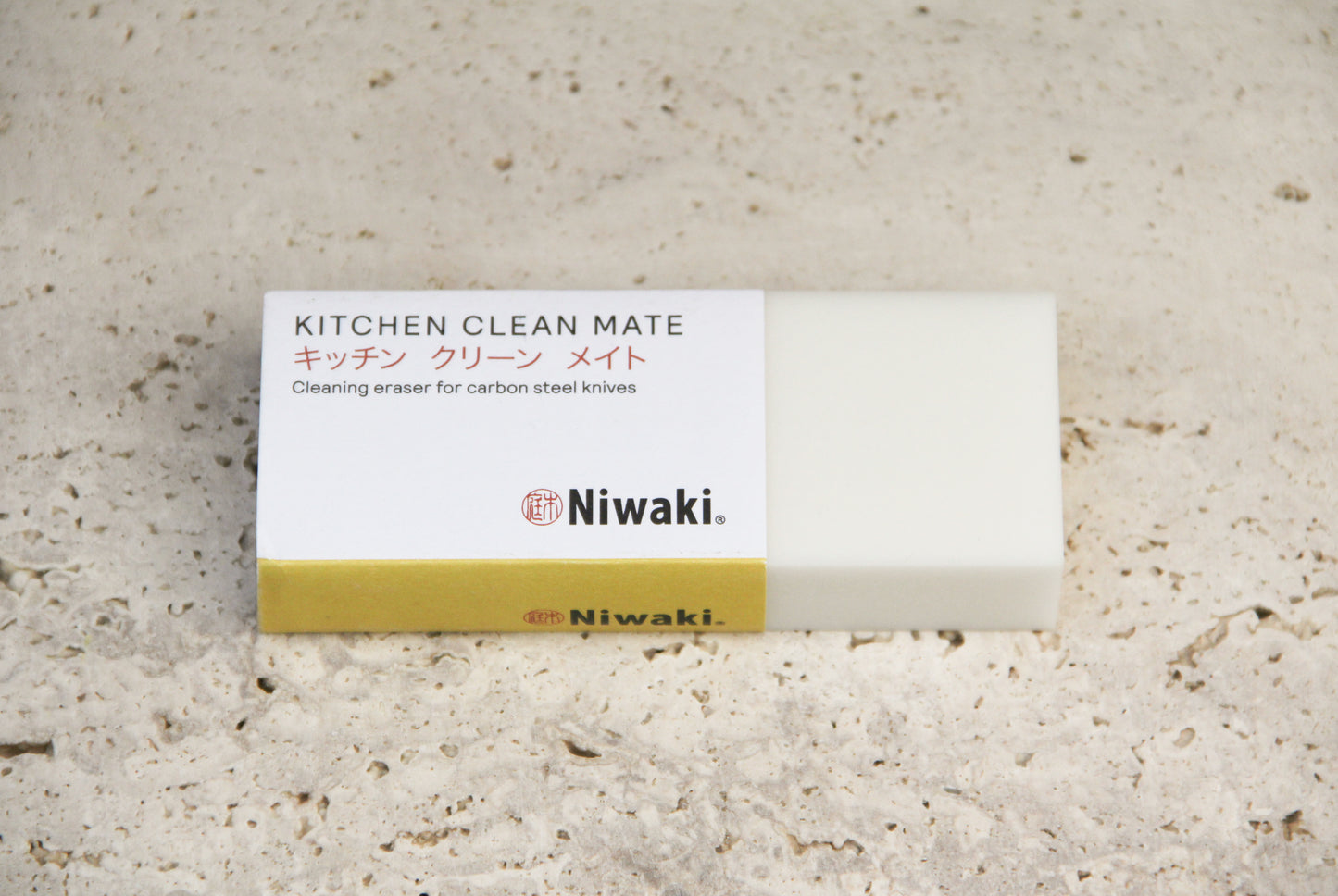 Ein rechteckiger, weiß-goldfarbener Radiergummi mit der Aufschrift „Niwaki | ‚Kitchen Clean Mate‘ Rostentferner für Küchenmesser von Niwaki“, ideal zum Reinigen von Messern aus Kohlenstoffstahl, liegt auf einer strukturierten, beigen Oberfläche.