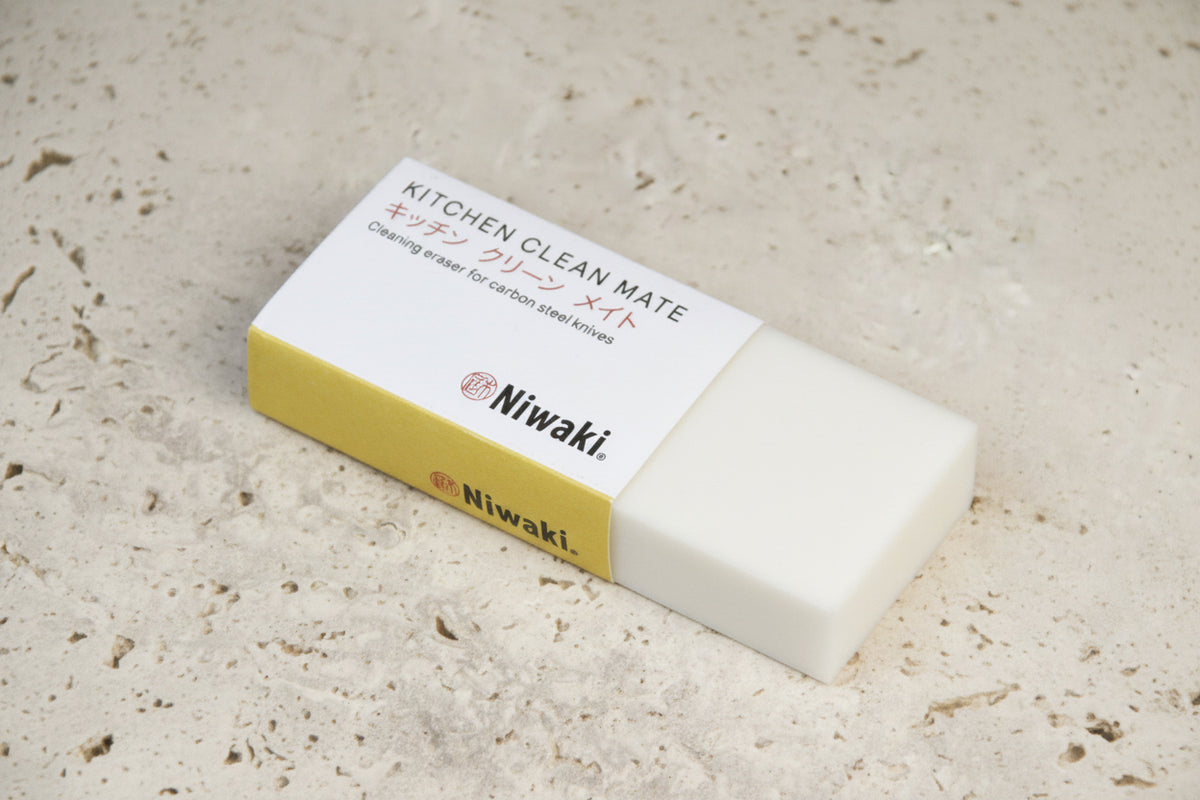 Ein rechteckiger, weiß-goldfarbener Radiergummi mit der Aufschrift „Niwaki | ‚Kitchen Clean Mate‘ Rostentferner für Küchenmesser von Niwaki“, ideal zum Reinigen von Messern aus Kohlenstoffstahl, liegt auf einer strukturierten, beigen Oberfläche.
