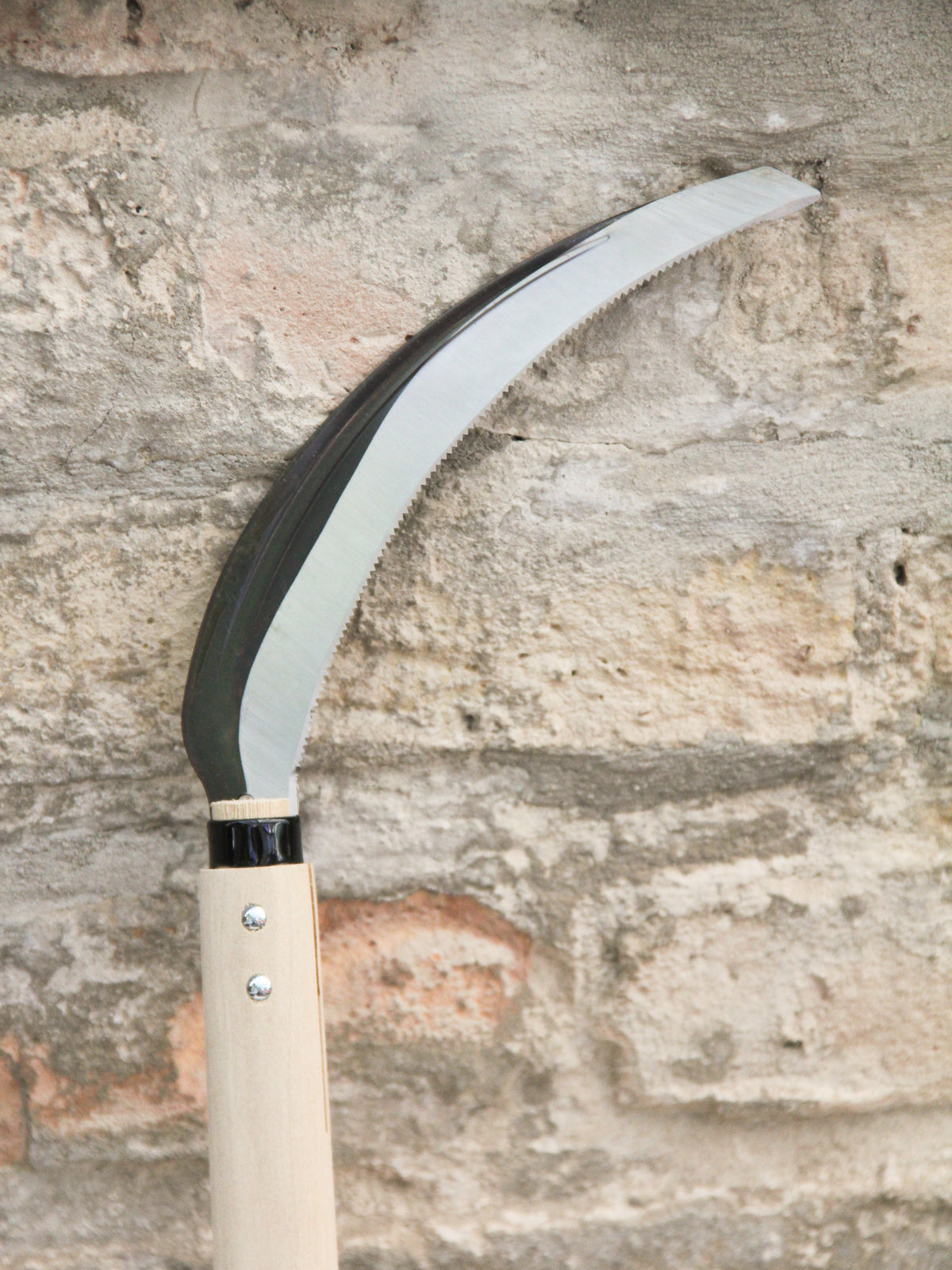 Eine Nahaufnahme einer Niwaki | „Herbaceous Sickle“ Nokogama Reissichel aus Kohlenstoffstahl, 32 cm – mit ihrer glänzenden, geschwungenen Klinge und dem hellen Holzgriff –, die an einer strukturierten Steinmauer lehnt.