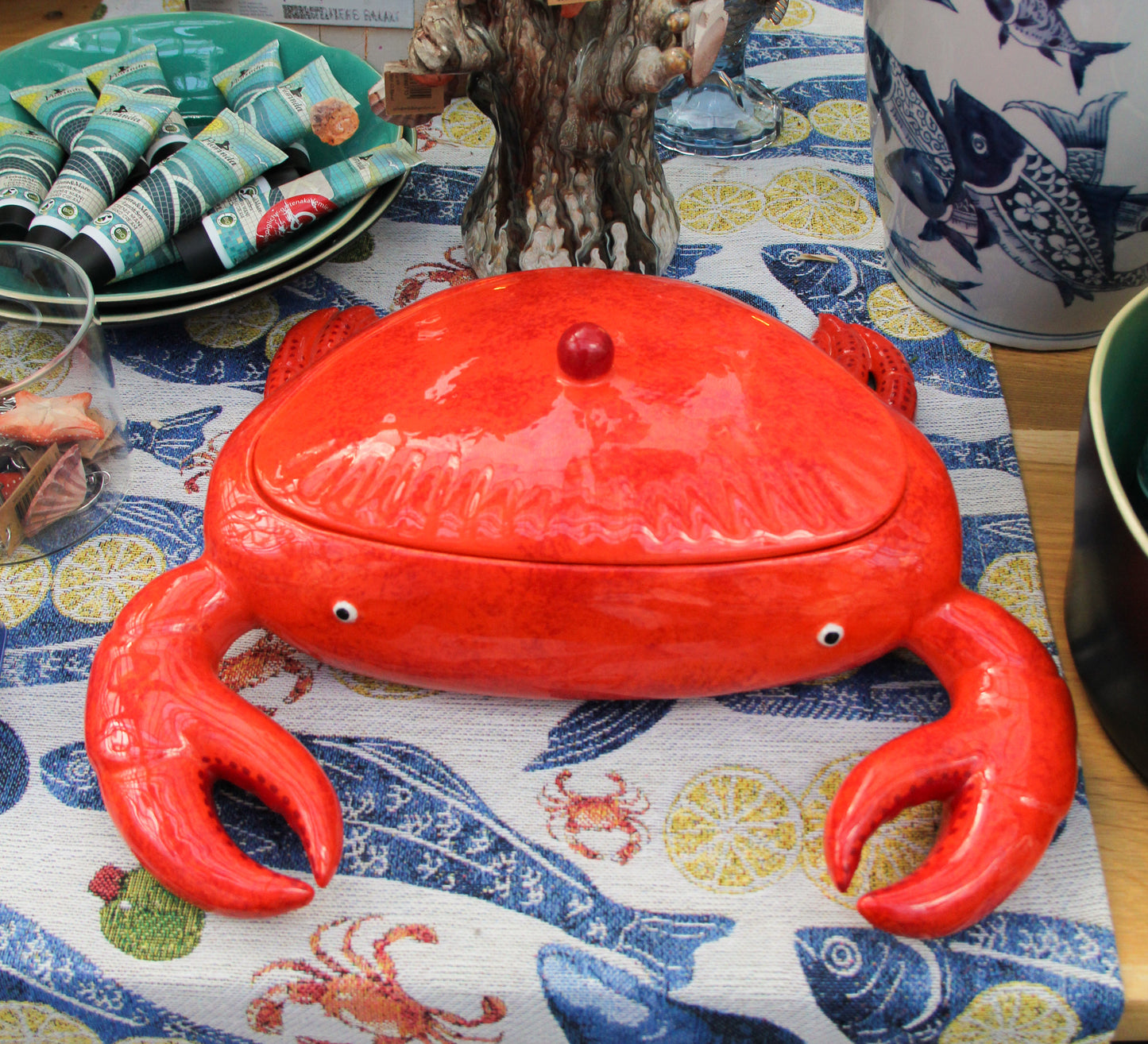 Eine leuchtend rote „Summer Bliss Crab“-Krabben-Schale von Costa Nova aus Keramik, die in Form einer Krabbe mit abnehmbarem Deckel gestaltet ist, steht auf einer Tischdecke mit maritimem Muster, umgeben von weiteren Dekorationselementen zum Thema Meeresfrüchte.
