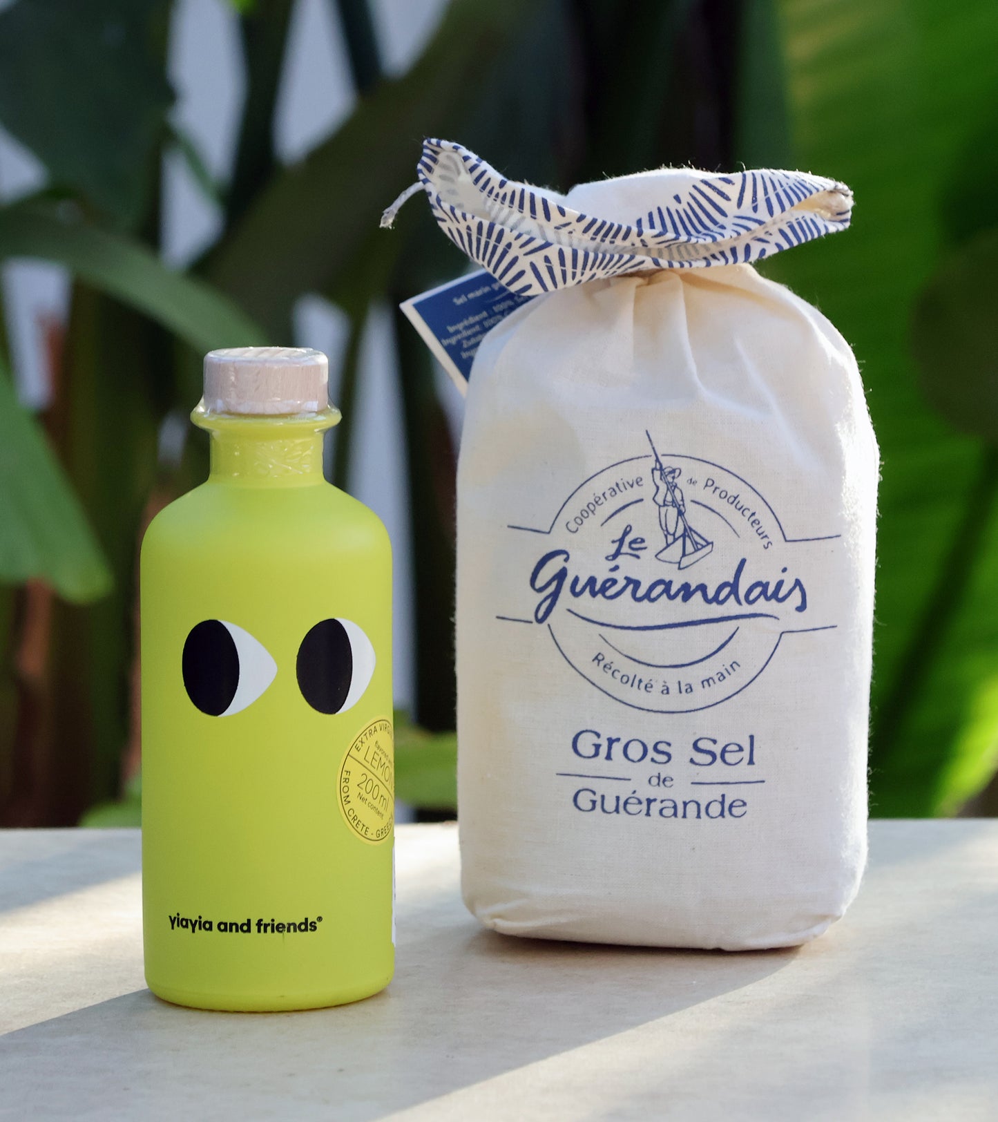 Eine grüne Flasche Yiayia and friends mit Cartoon-Augen steht neben einer Stofftasche mit Le Guérandais Guerandais | Grobes Meersalz aus der Bretagne, beide auf einer hellen Fläche mit sanft verschwommenen grünen Pflanzen im Hintergrund.