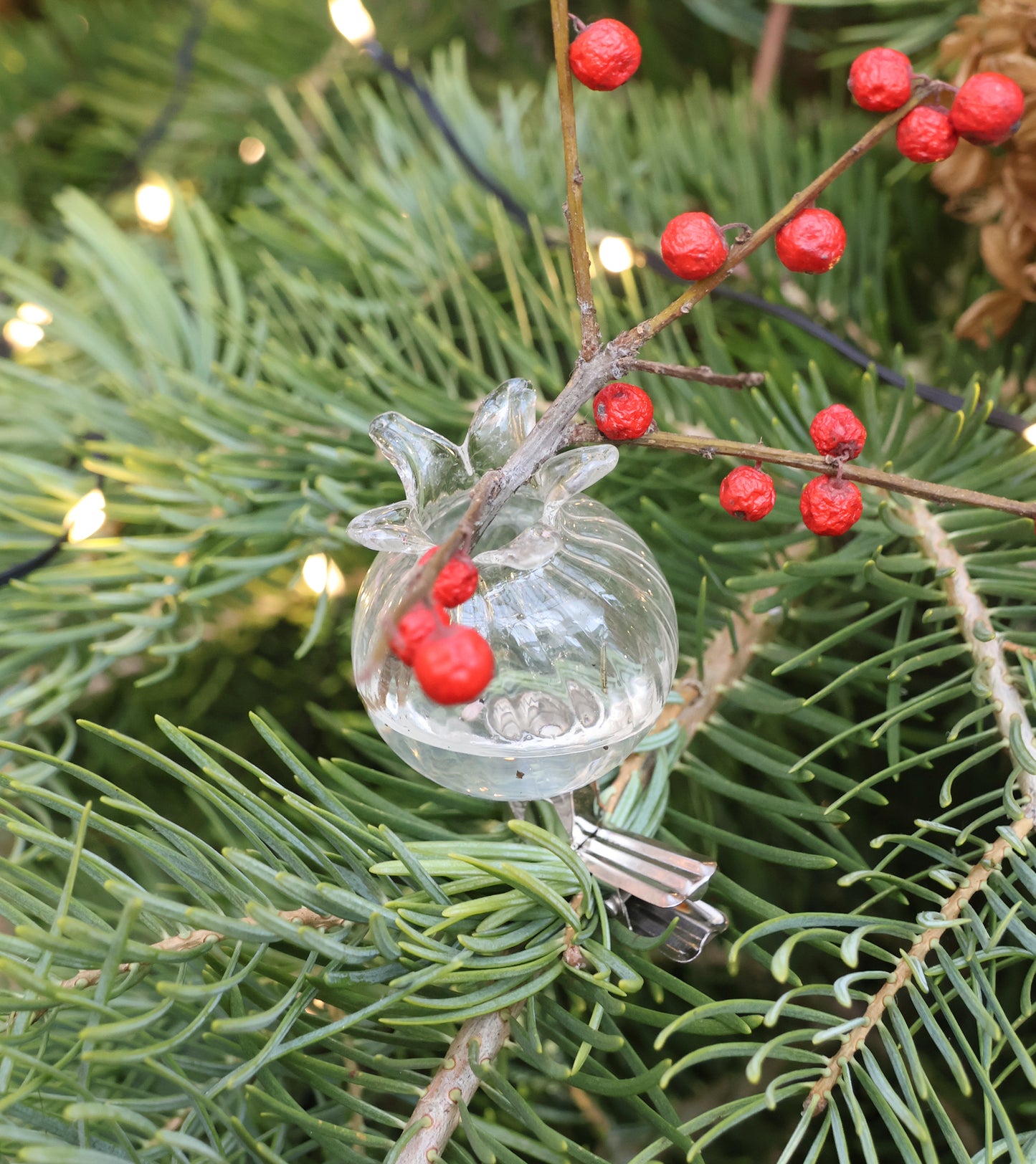 Ein Des Pots Filigrane Mini Klemmvasen aus dem 6er Set, gefüllt mit einem Zweig roter Beeren, wird auf die grünen Nadeln eines Weihnachtsbaums gesteckt, während im Hintergrund kleine weiße Lichter leuchten.
